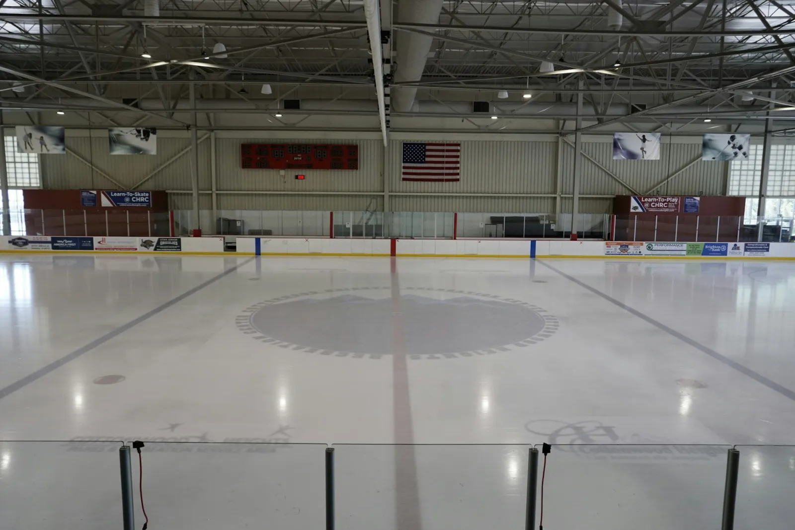 La pista vacía vista desde detrás del vidrio en un extremo, con una bandera grande y banderines rojos en la pared del fondo, bajo un techo de acero a la vista. — Cottonwood Heights Ice Arena