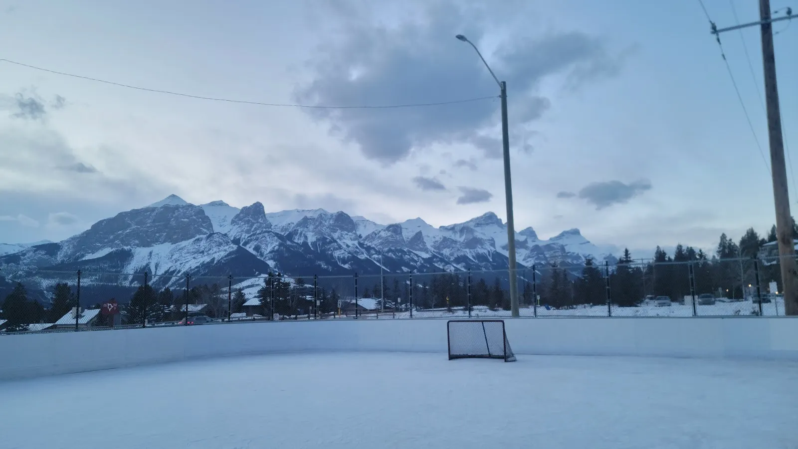 The boarded Cougar Creek rink in winter with a goal on the snowy ice, a basketball hoop behind and the mountains beyond. — Cougar Creek Boarded Ice Rink