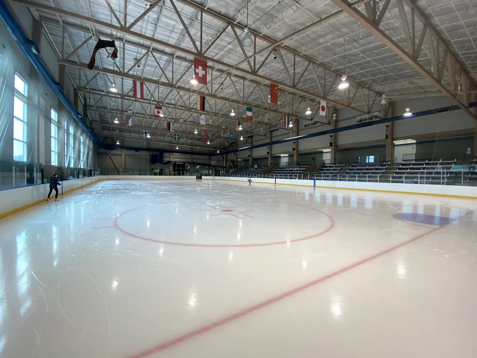 The full sheet from one corner, with national flags hanging from the roof trusses, tall windows along the left wall and bleachers on the right. — County Ice Center
