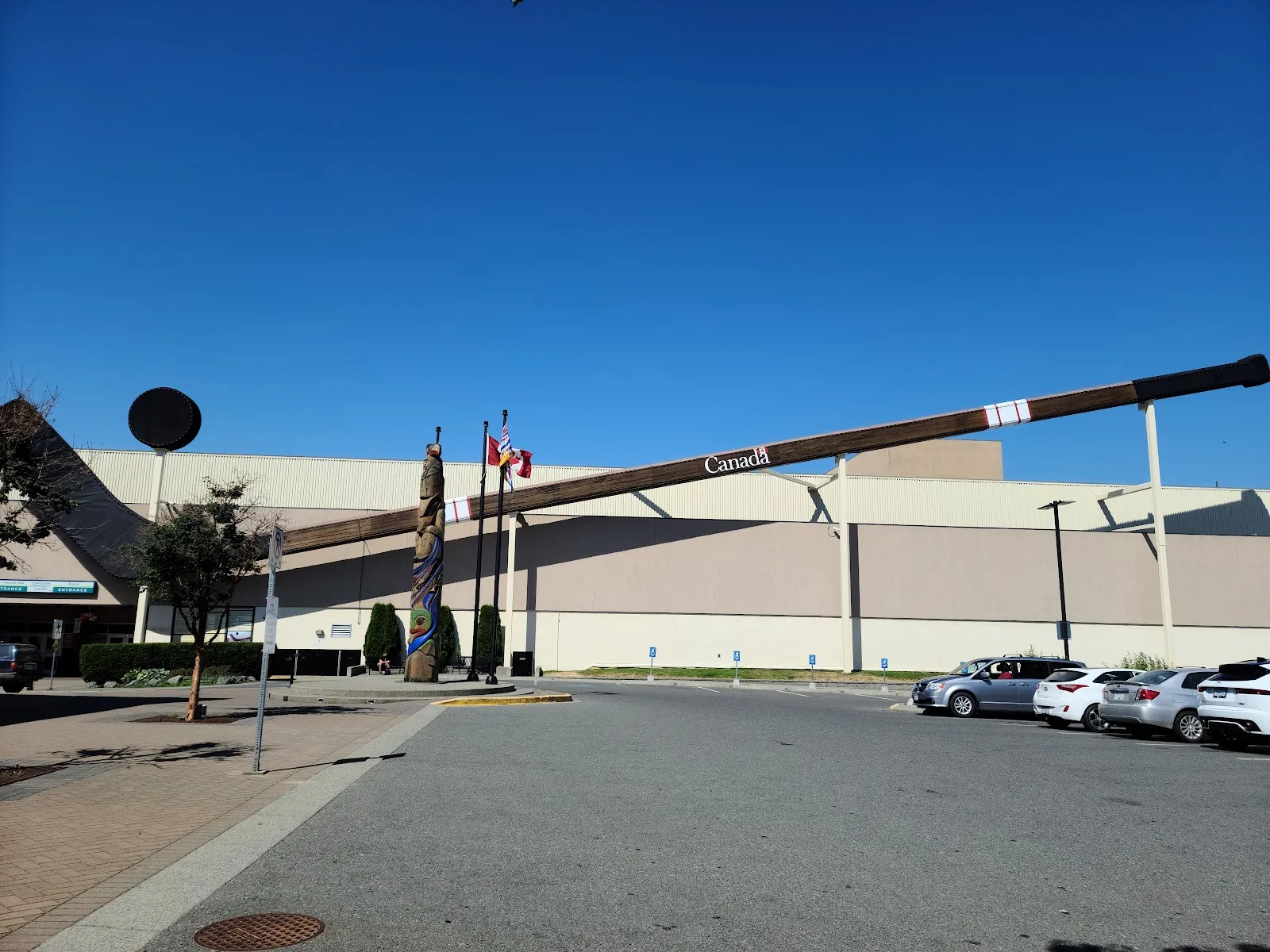 The wide beige wall of the Cowichan arena building with the giant hockey stick marked Canada mounted across its front, a carved pole and flags in the plaza and cars parked in front under a deep blue sky. — Cowichan Arena (Cowichan Community Centre)