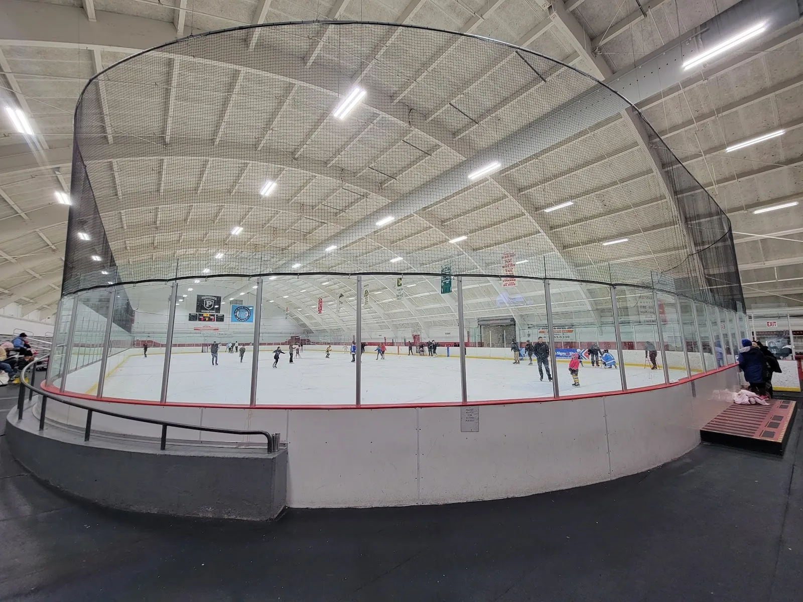 A wide fisheye view of the sheet inside the white dome, skaters spread across the ice behind a curved glass-topped rail. — Cranston Veterans Memorial Ice Rink