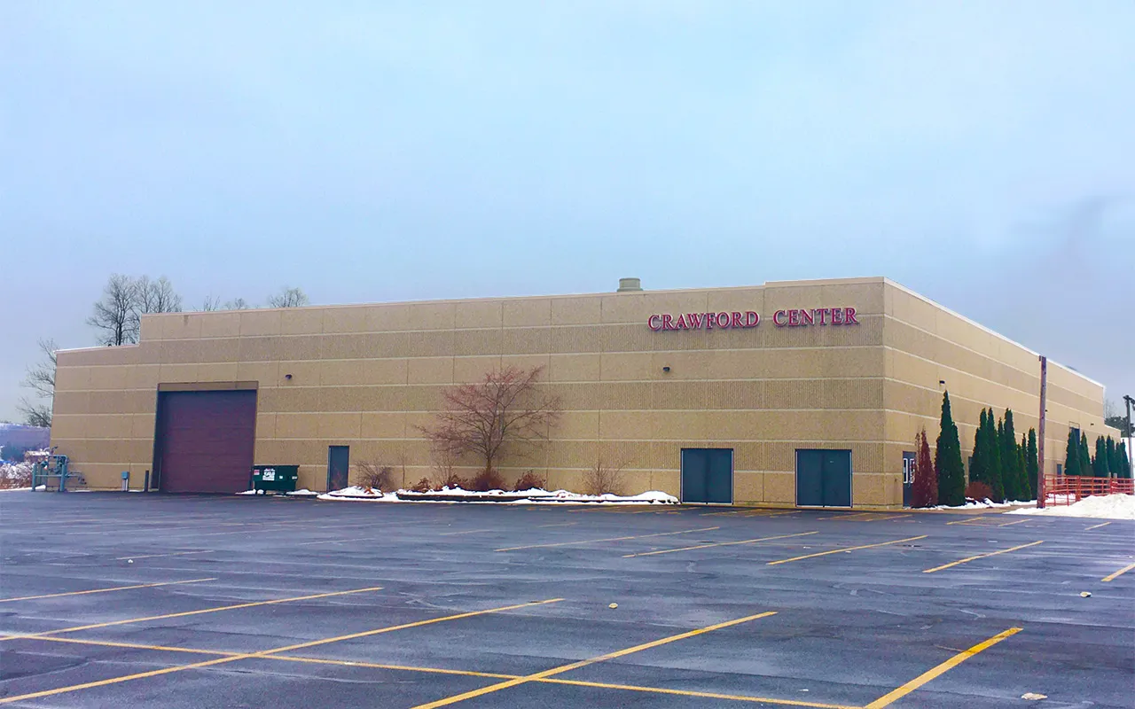The beige Crawford Center building with its name on the upper wall, a large overhead door and a car park in front, under a pale winter sky. — Crawford Center