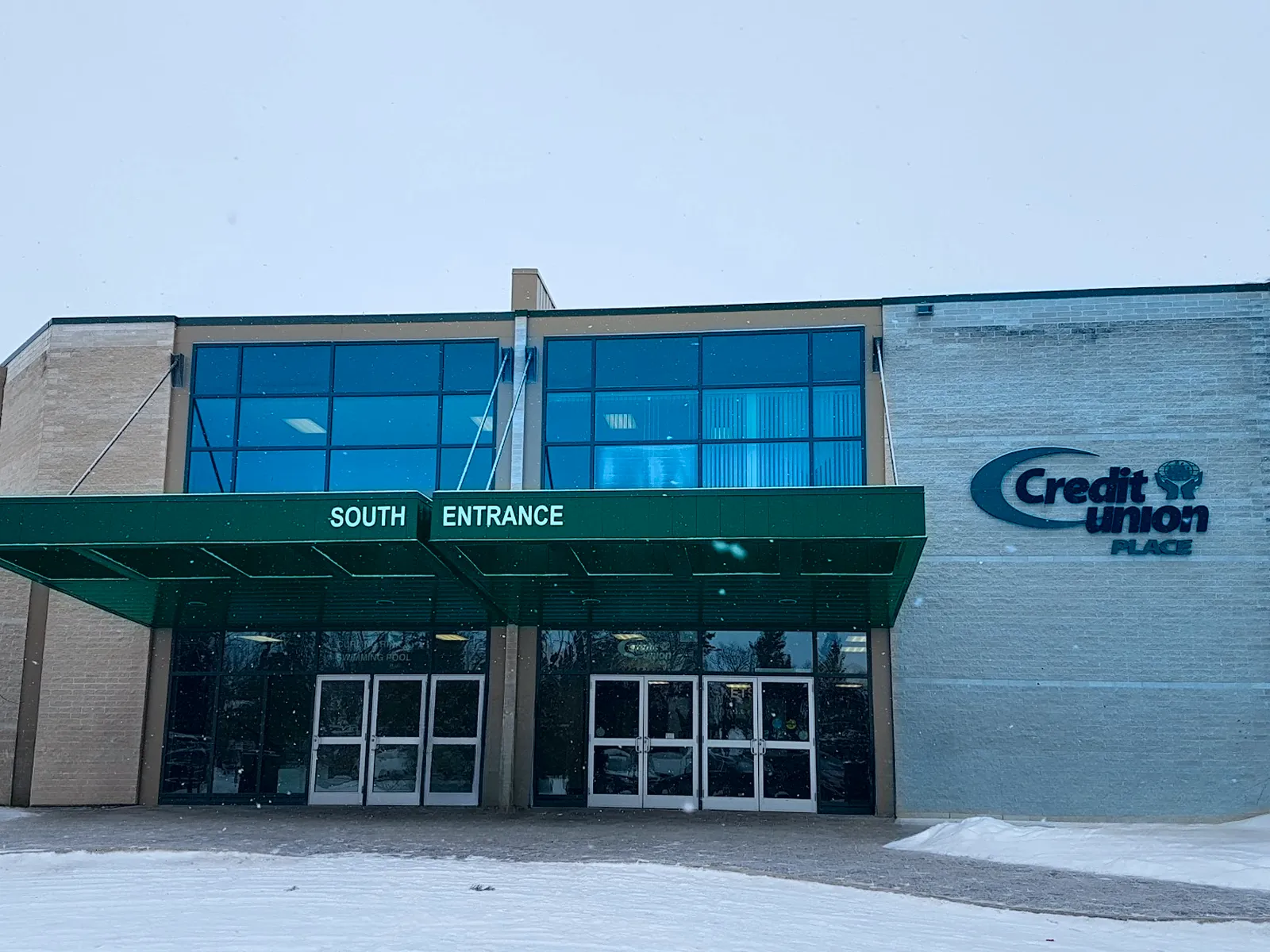 The south entrance of Credit Union Place in winter: glass doors under a green canopy marked South Entrance, with the Credit Union Place sign on the brick wall — Credit Union Place (Dauphin)