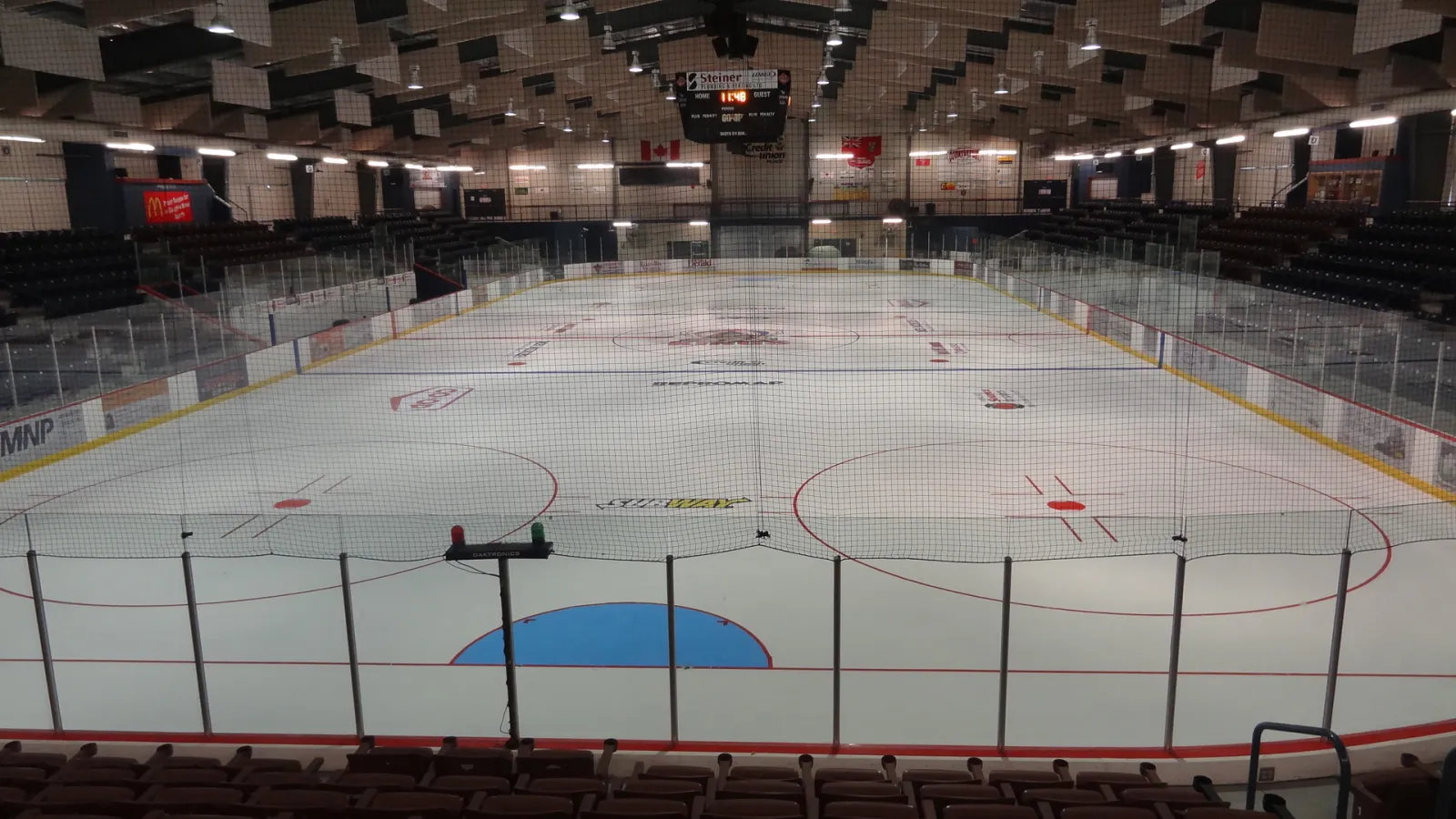 The empty Credit Union Place ice from the end seats behind the netting, with the Dauphin Kings logo at centre ice, the C-bowl seating and the scoreboard — Credit Union Place (Dauphin)