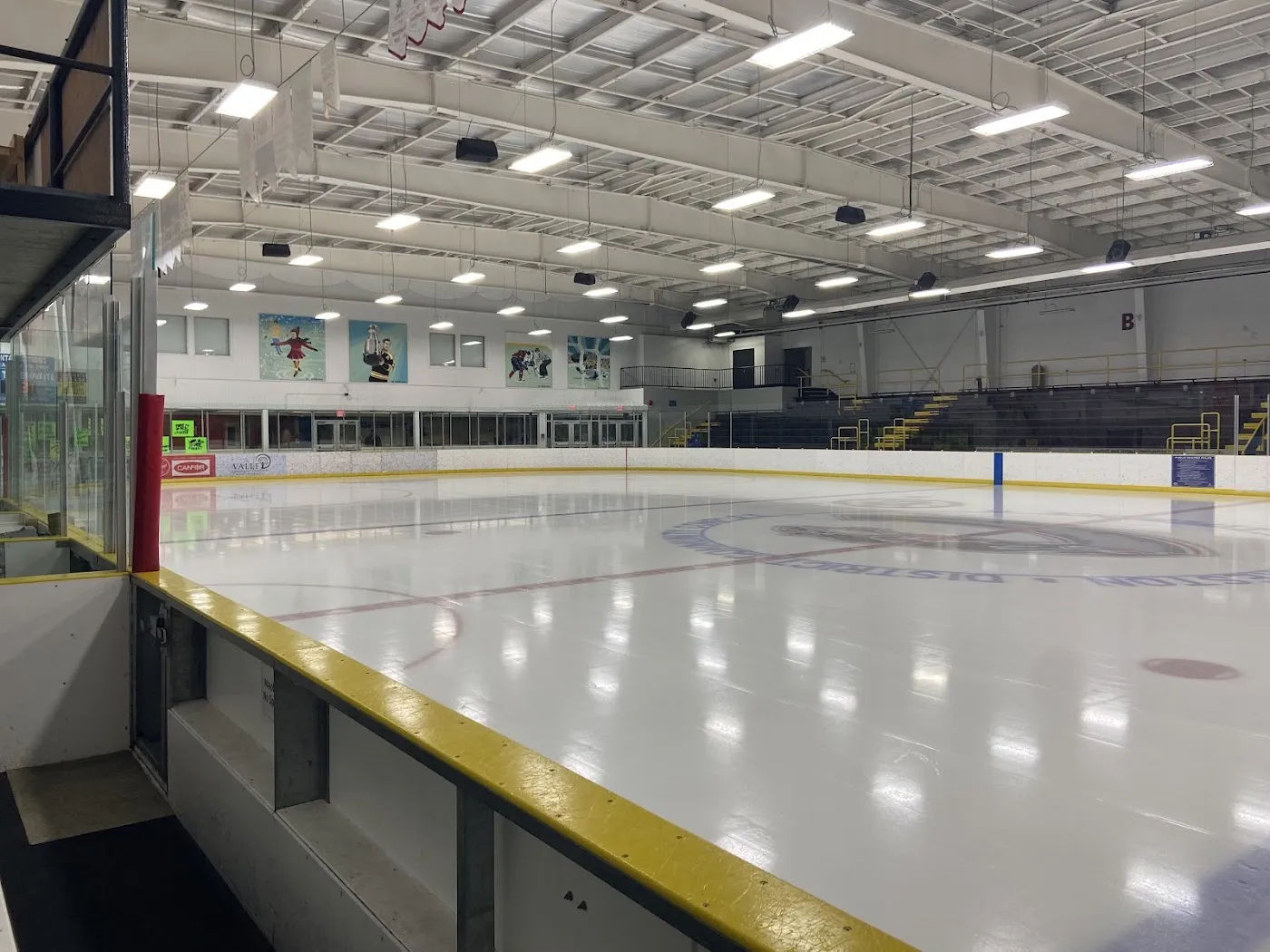 The empty ice sheet seen along the boards, with yellow kickplates, the far bleachers and a mural-covered end wall. — Creston & District Community Complex