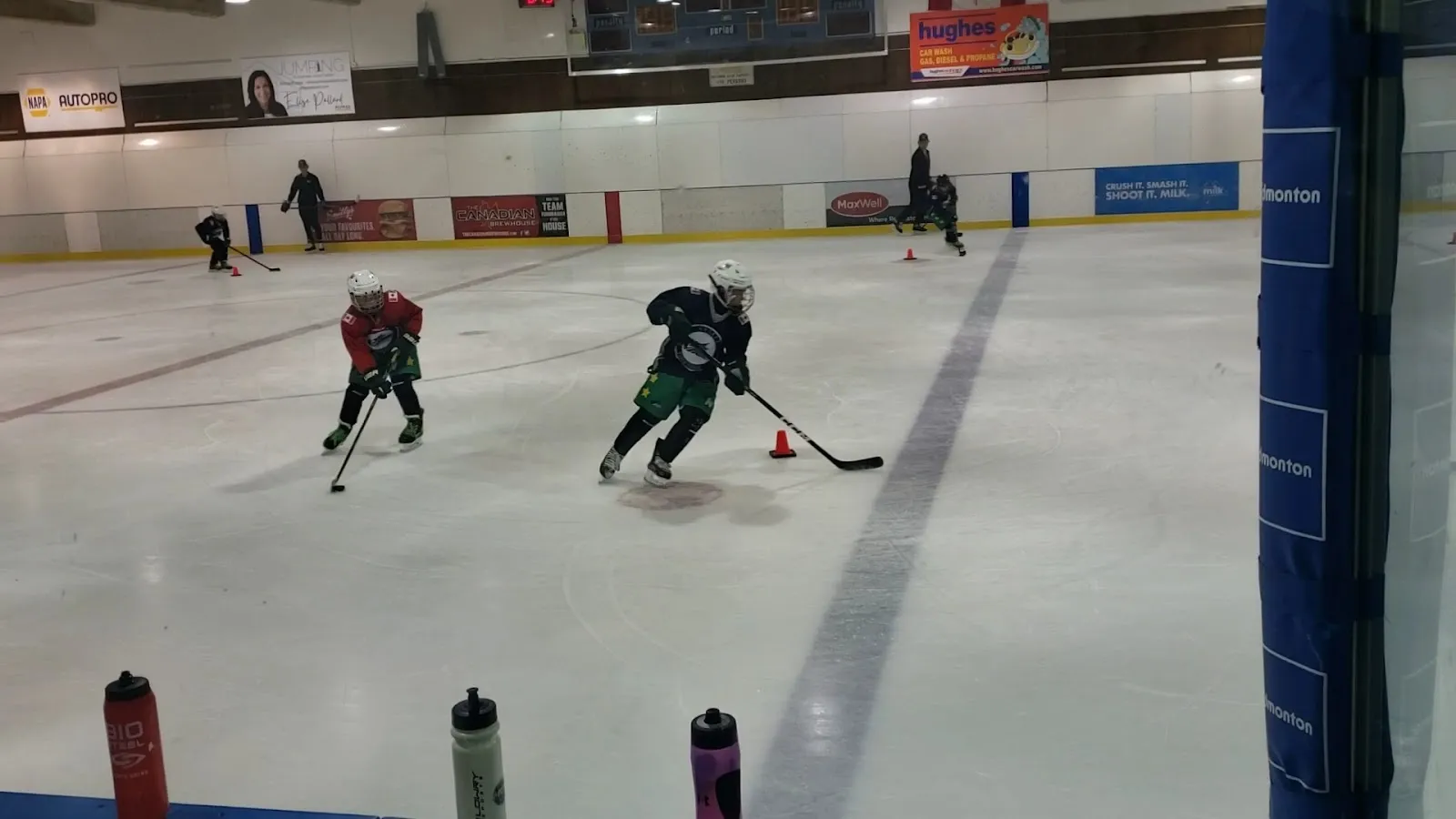 Two players skating a drill across a wide sheet, blue and white sponsor boards and a bright ceiling behind. — Crestwood Arena