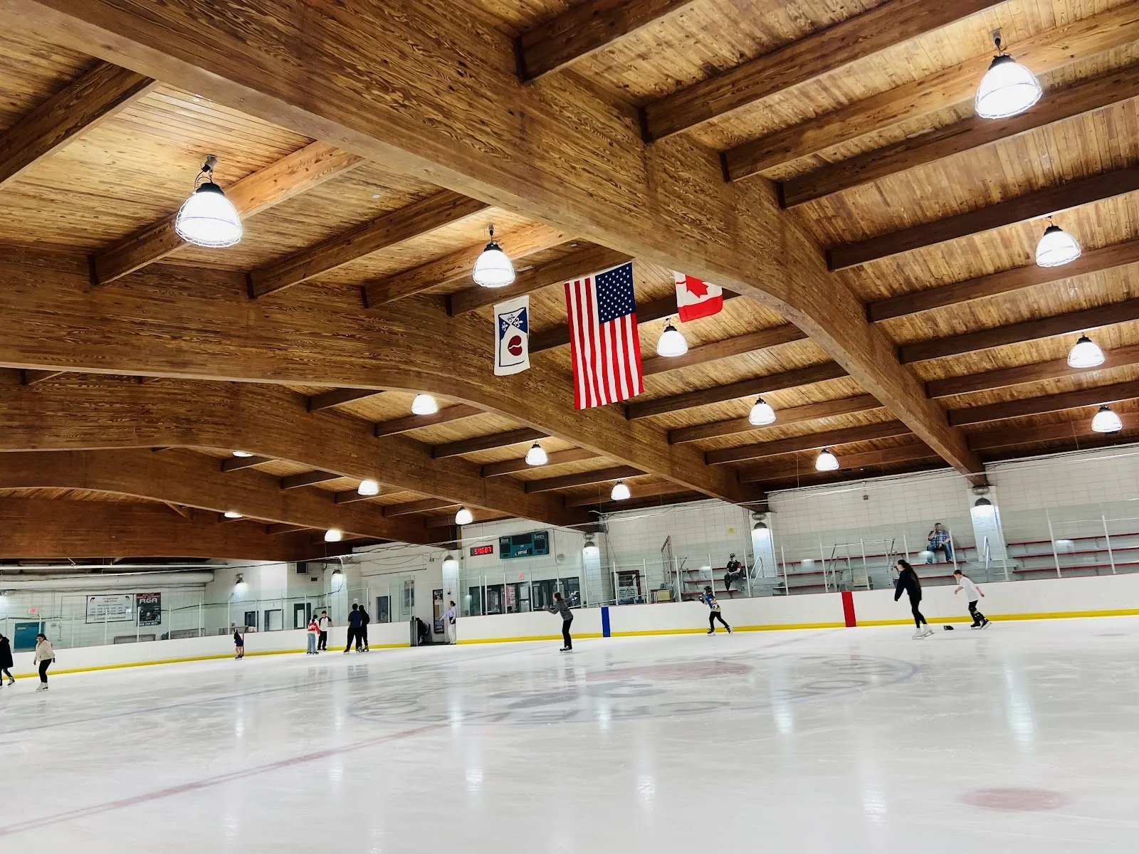 Una sesión pública en una pista amplia, con patinadores repartidos a lo largo de las vallas del fondo bajo un techo de madera con banderas colgadas de las vigas y estandartes con la hoja de maple canadiense. — Creve Coeur Ice Arena