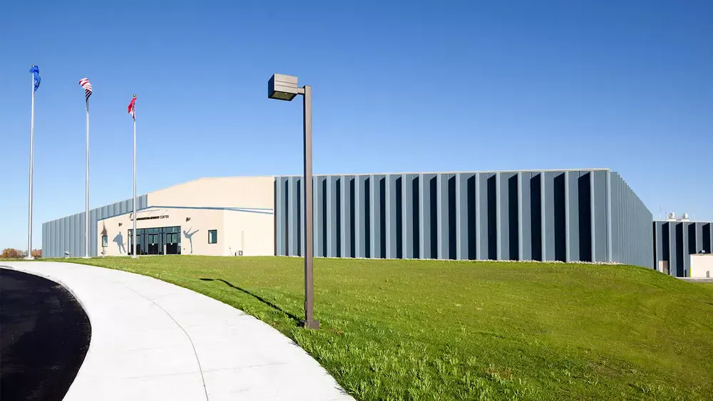 The Crookston Sports Center from the curving front walk: a tan entrance block with skater silhouettes, flagpoles, and the long blue-gray ribbed wall of the arenas behind a grassy berm. — Crookston Sports Center