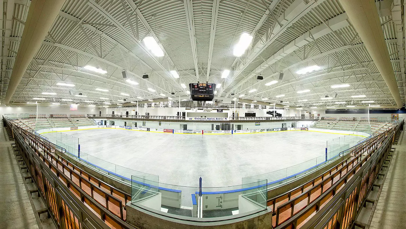 A wide view of the empty main arena from the wooden seats, with the center-hung scoreboard under white steel trusses. — Crookston Sports Center