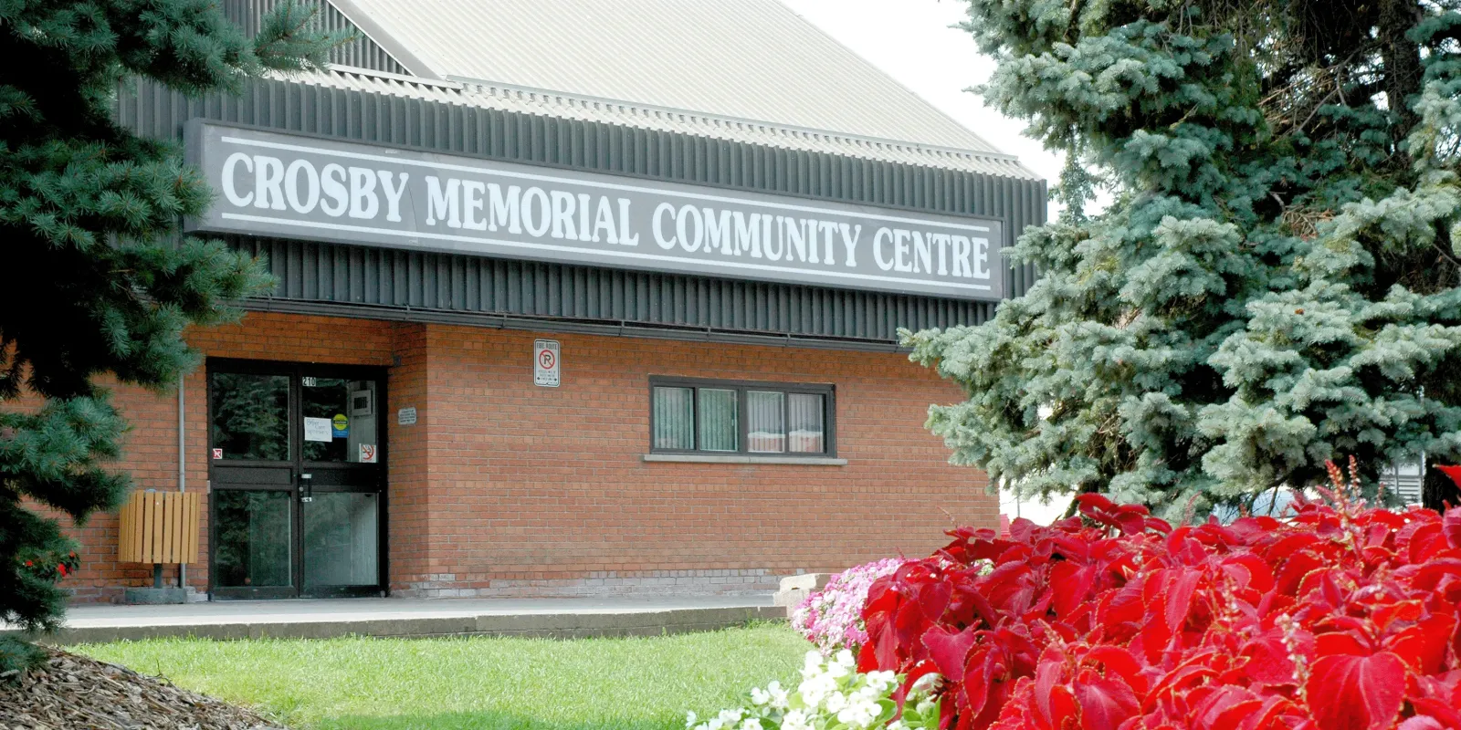 Entrada principal de ladrillo del Crosby Memorial Community Centre, con el letrero iluminado del edificio sobre la puerta y arbustos al frente. — Crosby Community Centre