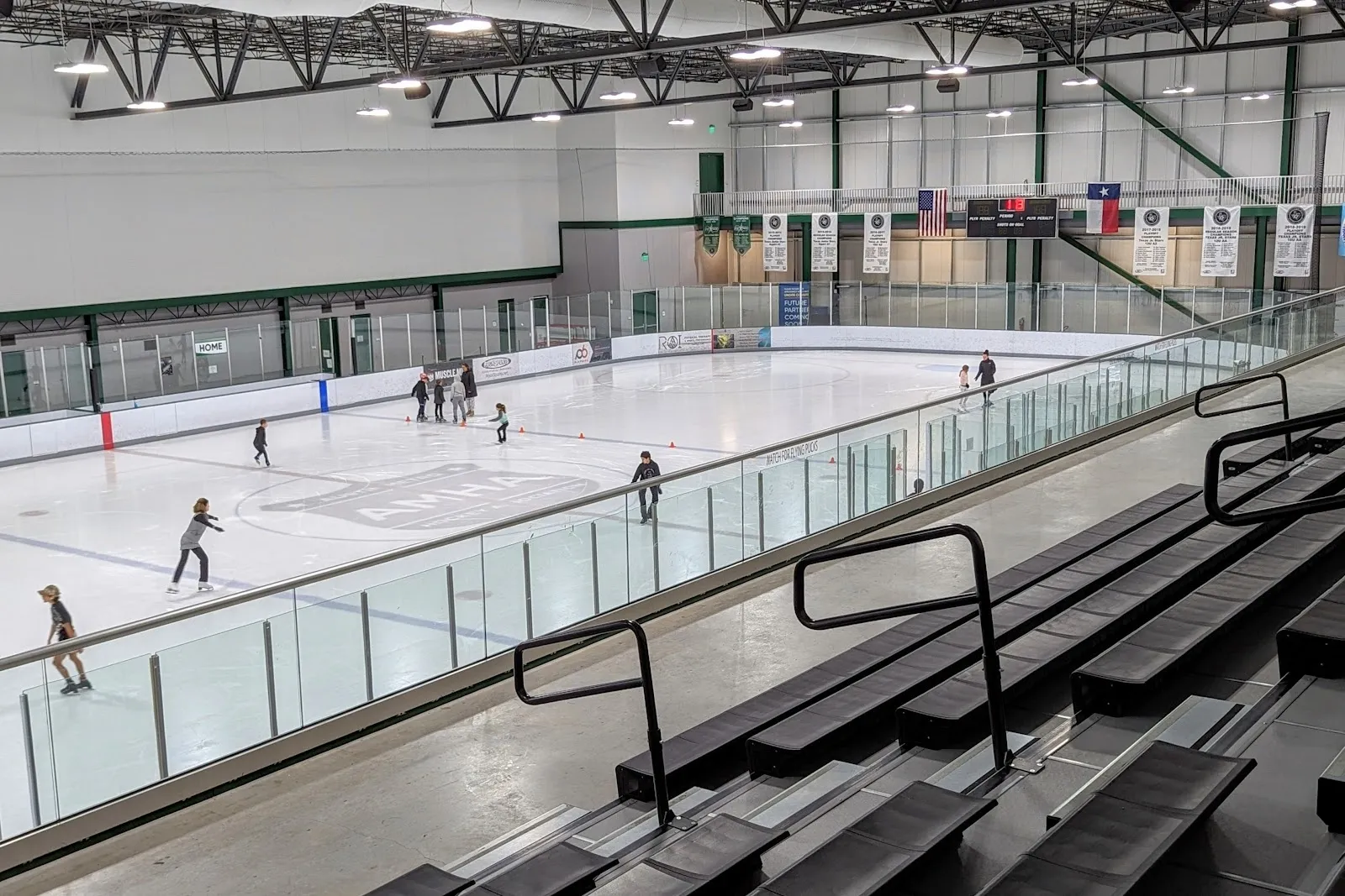 Una pista amplia vista desde las gradas, con patinadores y jugadores repartidos por el hielo blanco detrás del vidrio de la pista, en un recinto luminoso con estructura de acero. — Ice & Field at the Crossover