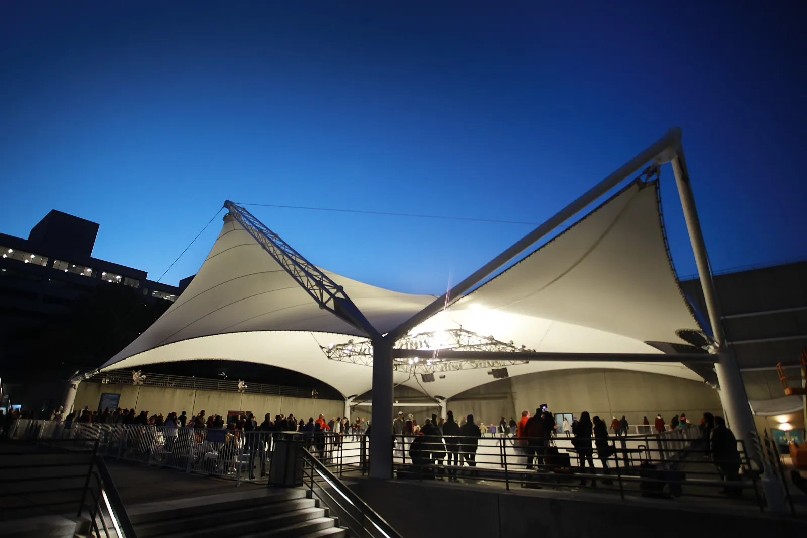 El techo blanco de tela tensada de Ice Terrace en Crown Center al atardecer, iluminado desde dentro, con curiosos a lo largo de la baranda — Crown Center Ice Terrace