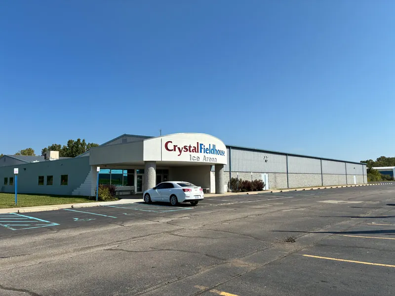 Frente del edificio metálico blanco y bajo con una marquesina de entrada redondeada que dice "Crystal Fieldhouse Ice Arena", con el estacionamiento al frente bajo un cielo despejado — Crystal Fieldhouse