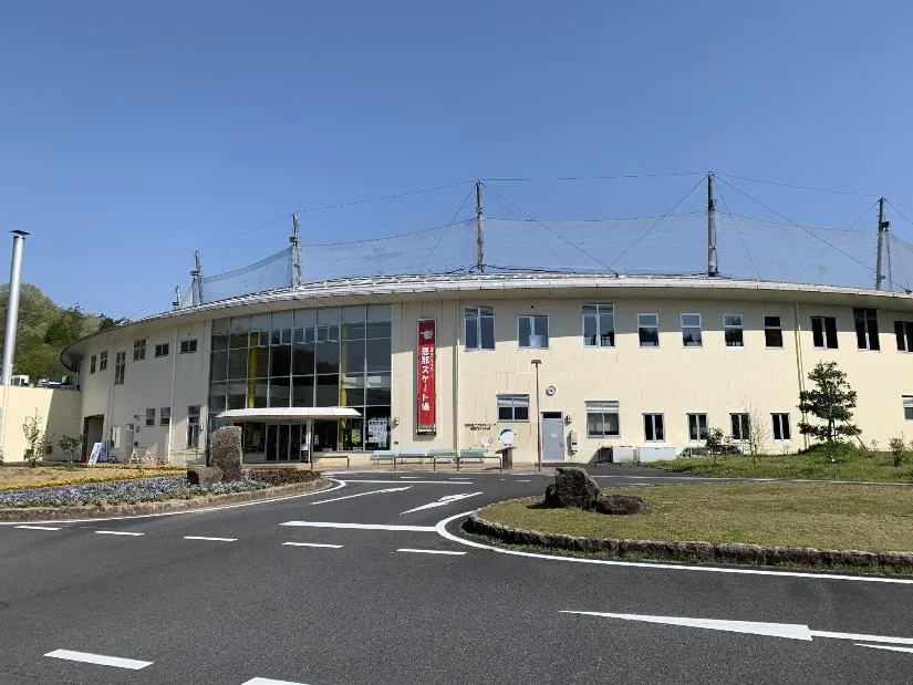 晴れた空の下、駐車場から見た丸みのあるクリーム色のセンターハウス。ガラス張りの入口、「恵那スケート場」と書かれた赤い垂れ幕、屋根の上のネットが見えます。 — Gifu Crystal Park Ena Skate Rink