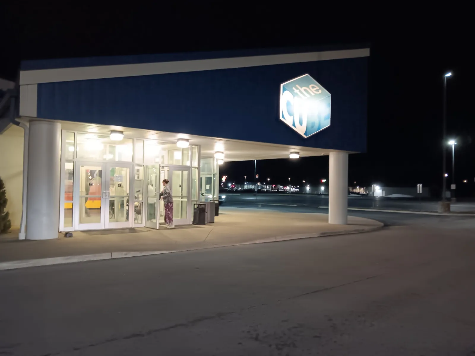 La entrada de vidrio de noche bajo una marquesina iluminada, con el logotipo de The Cube en la fachada azul arriba. — The Cube Ice Arena