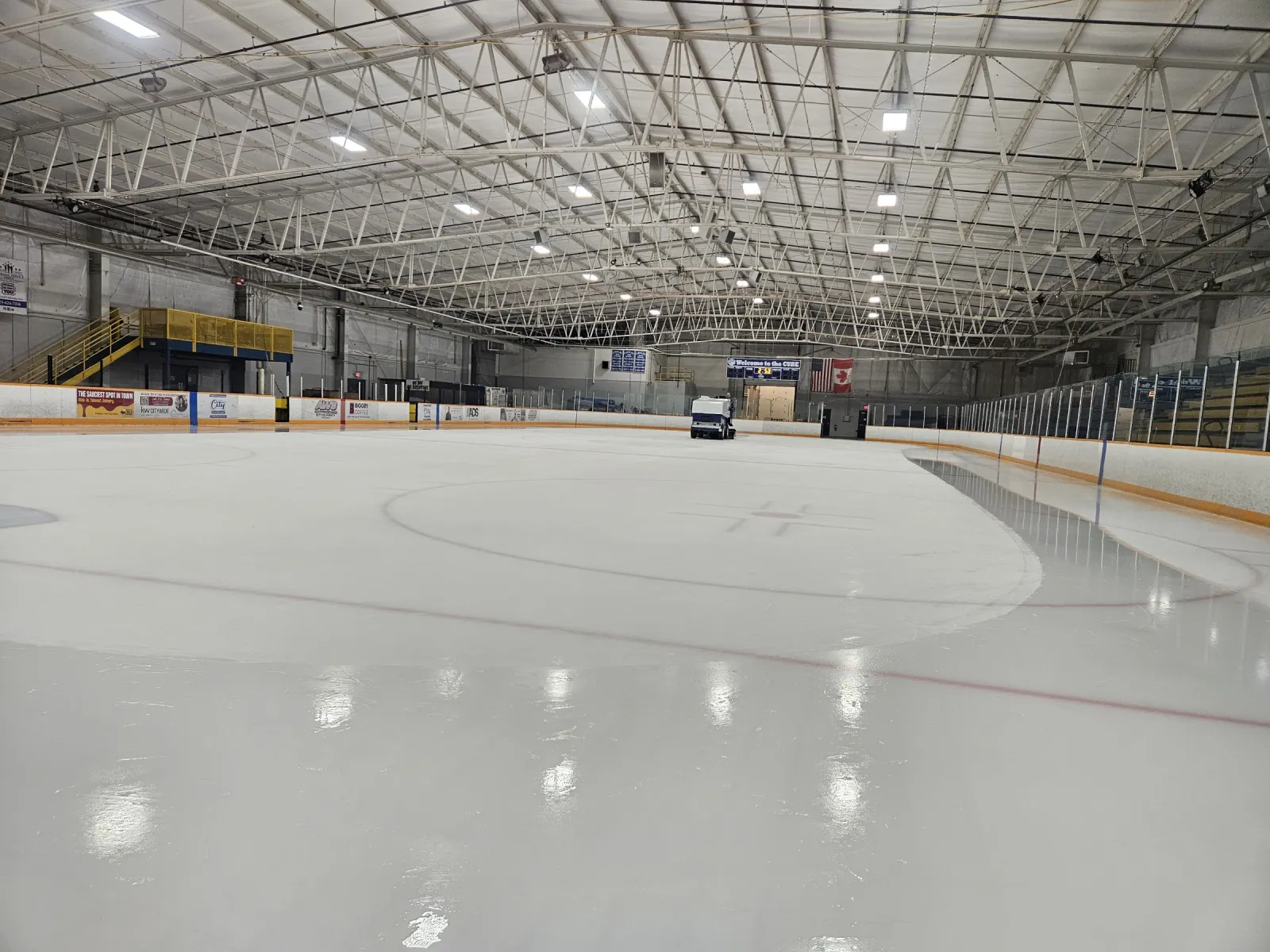 Una pista de hielo vacía bajo un techo blanco de armaduras de acero, con vallas de borde naranja y el cristal a ambos lados. — The Cube Ice Arena