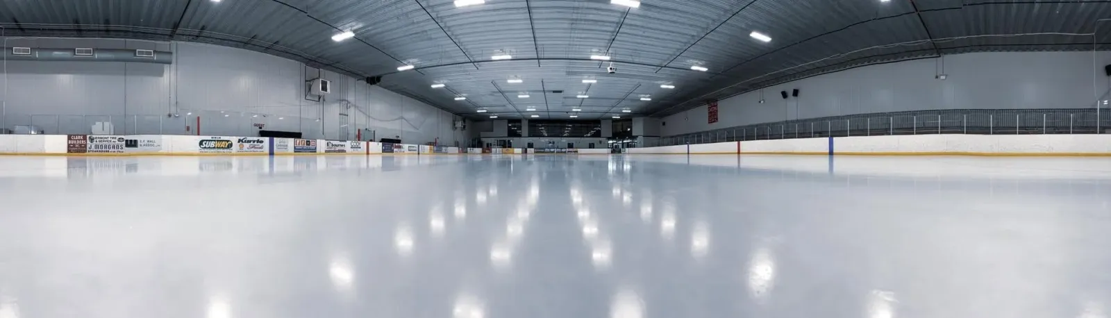 Wide low-angle view down the length of the empty ice sheet toward the far end boards, under a curved corrugated-metal roof with rows of ceiling lights; dasherboard ads (Subway, Vermont Tire & Service, J. Morgan's Steakhouse, Kurrie Foods and others) line the boards on both sides, no people or skaters present. — Central Vermont Memorial Civic Center