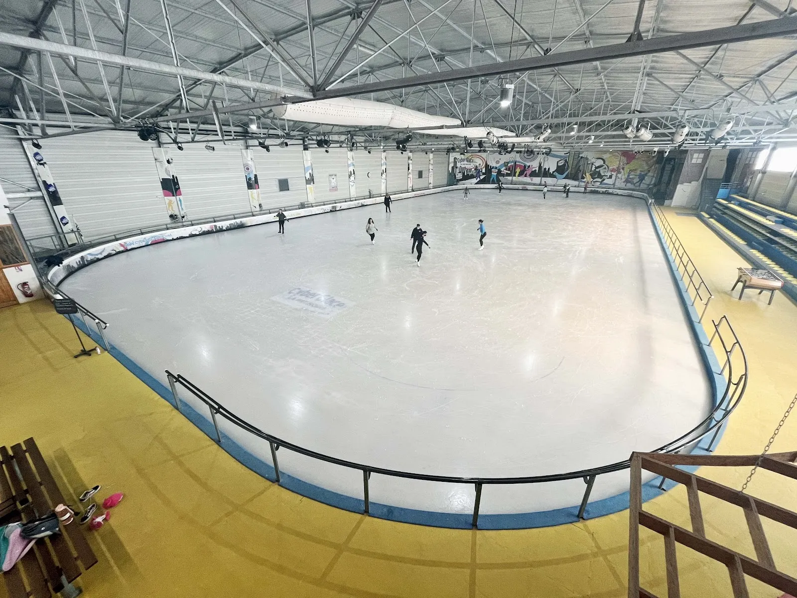 La piste de 56 sur 26 m vue des gradins sous la toiture à fermes d’acier, avec les bandes arrondies, le sol jaune autour de la patinoire et quelques patineurs sur la glace — Patinoire CyberGlace