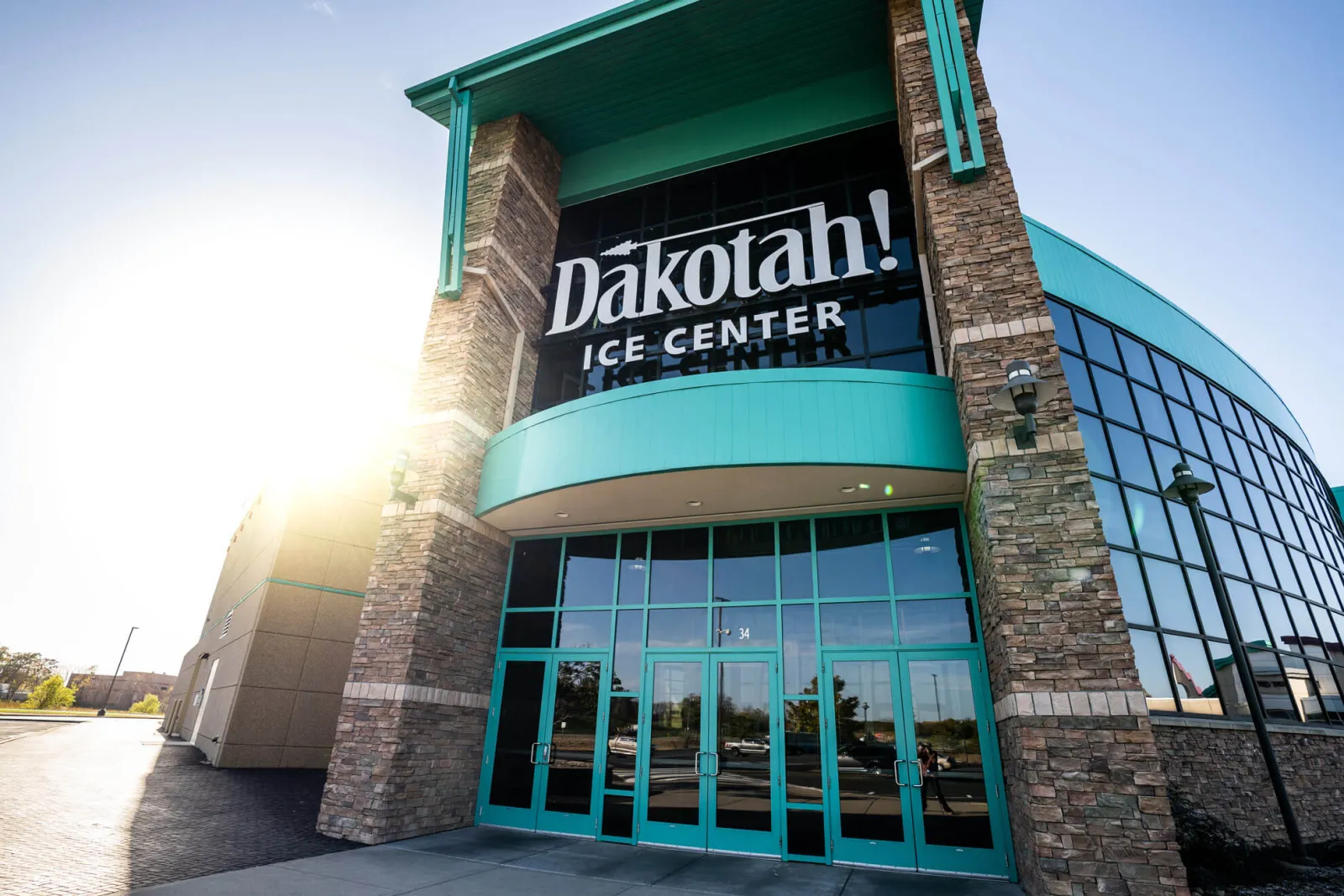 The Dakotah! Ice Center entrance: a teal canopy and glass doors between stone pillars, with a curved glass wall to the right. — Dakotah! Ice Center