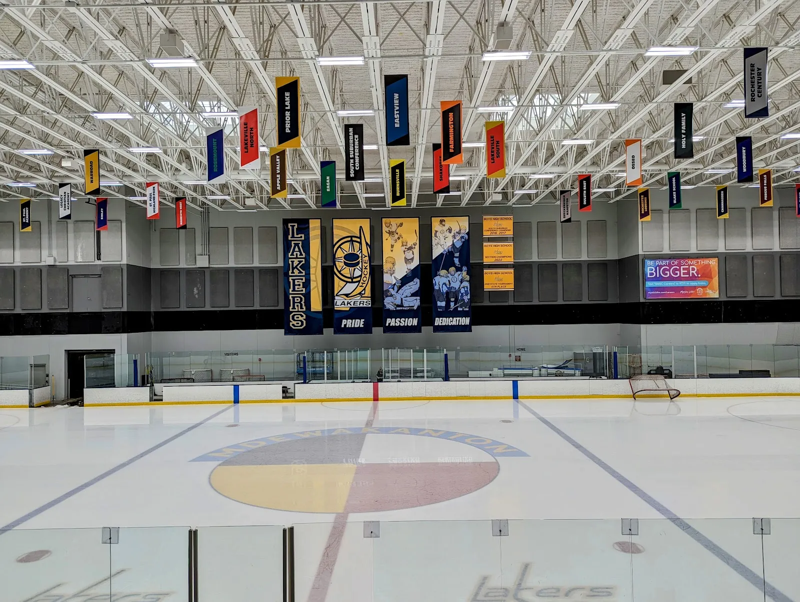 An empty sheet seen from behind the glass, with a four-colour circle at centre ice, Lakers banners on the far wall and team pennants hanging from the roof trusses. — Dakotah! Ice Center