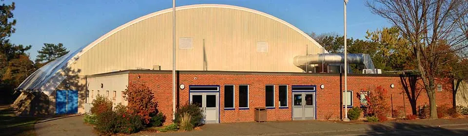Entrada principal de Daly Memorial Rink, un edificio de pista con techo de bóveda de cañón, base de ladrillo y entrada con puertas de vidrio, fotografiado de día desde el estacionamiento. — Daly Memorial Rink