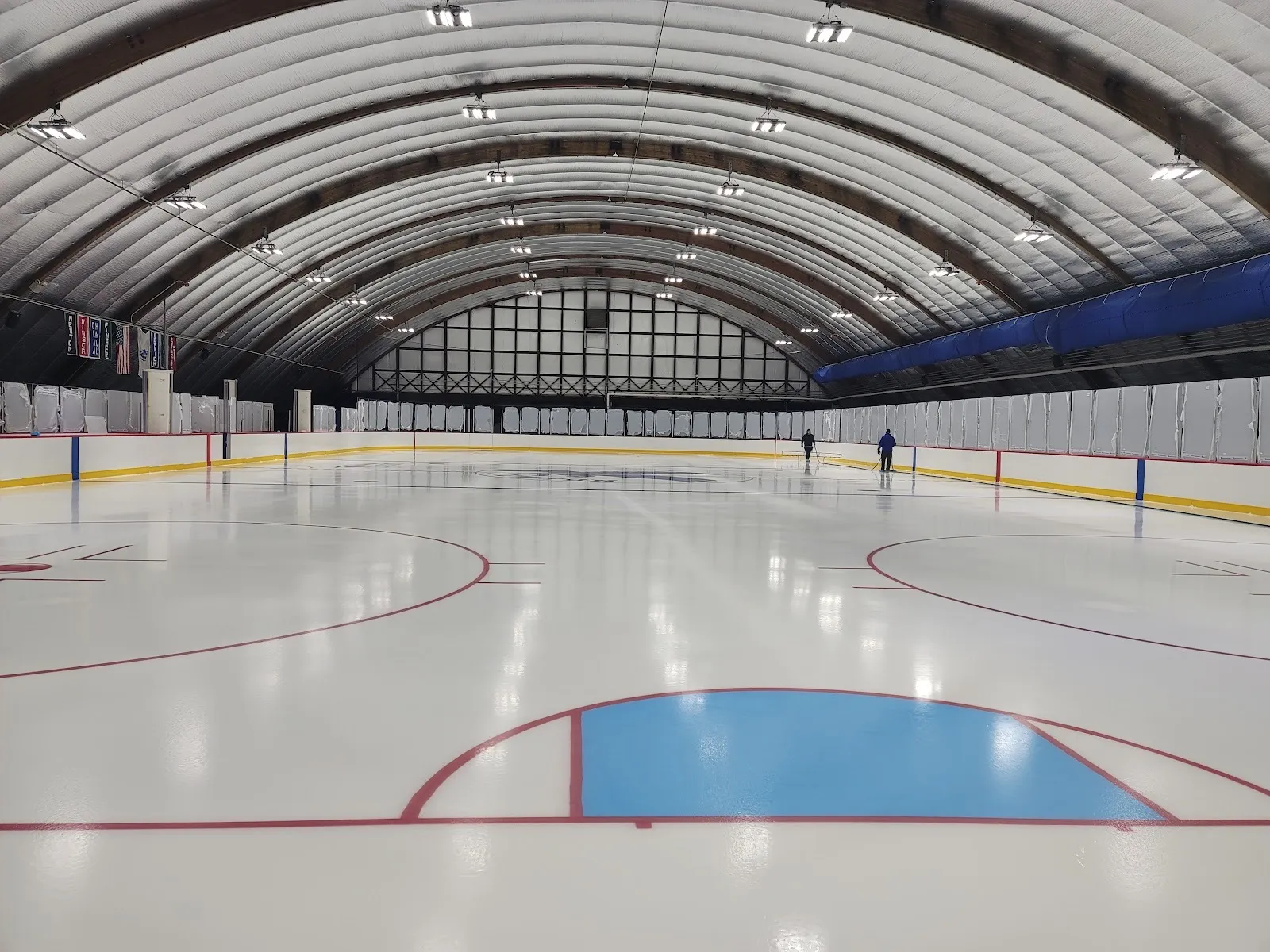 Toda la pista de Daly bajo los arcos curvos de madera del techo, recién pintada, con el área azul del portero al frente y el muro del fondo con ventanas a lo lejos — Daly Memorial Rink