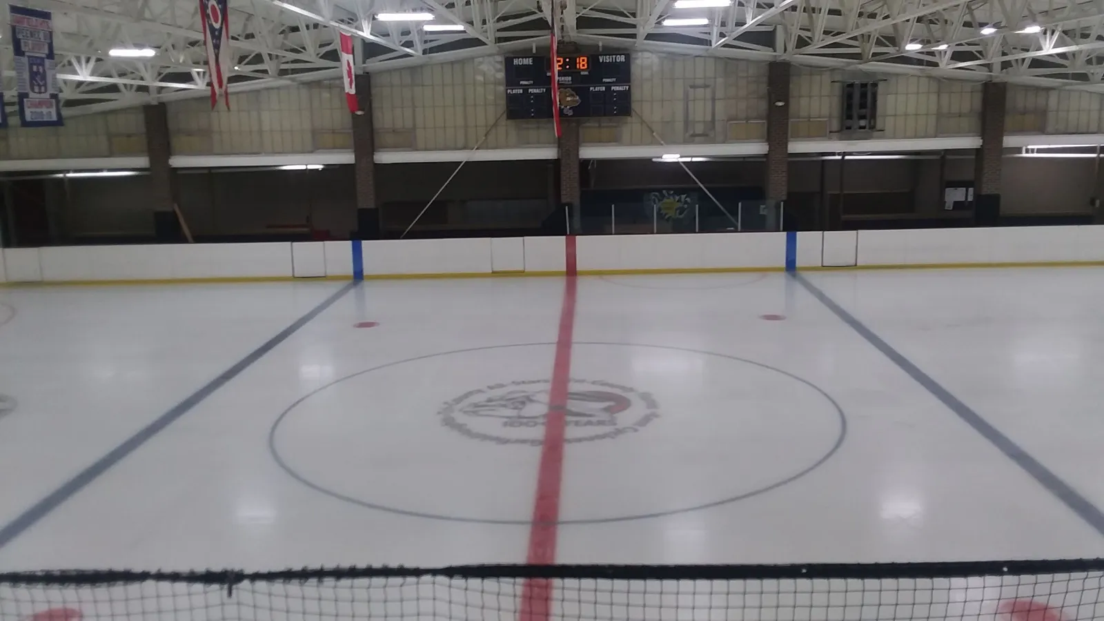 The empty ice at center line, seen through the end netting, with the Home and Visitor scoreboard, Ohio and Canadian flags and championship banners under the steel roof trusses. — Dan Kostel Recreation Center