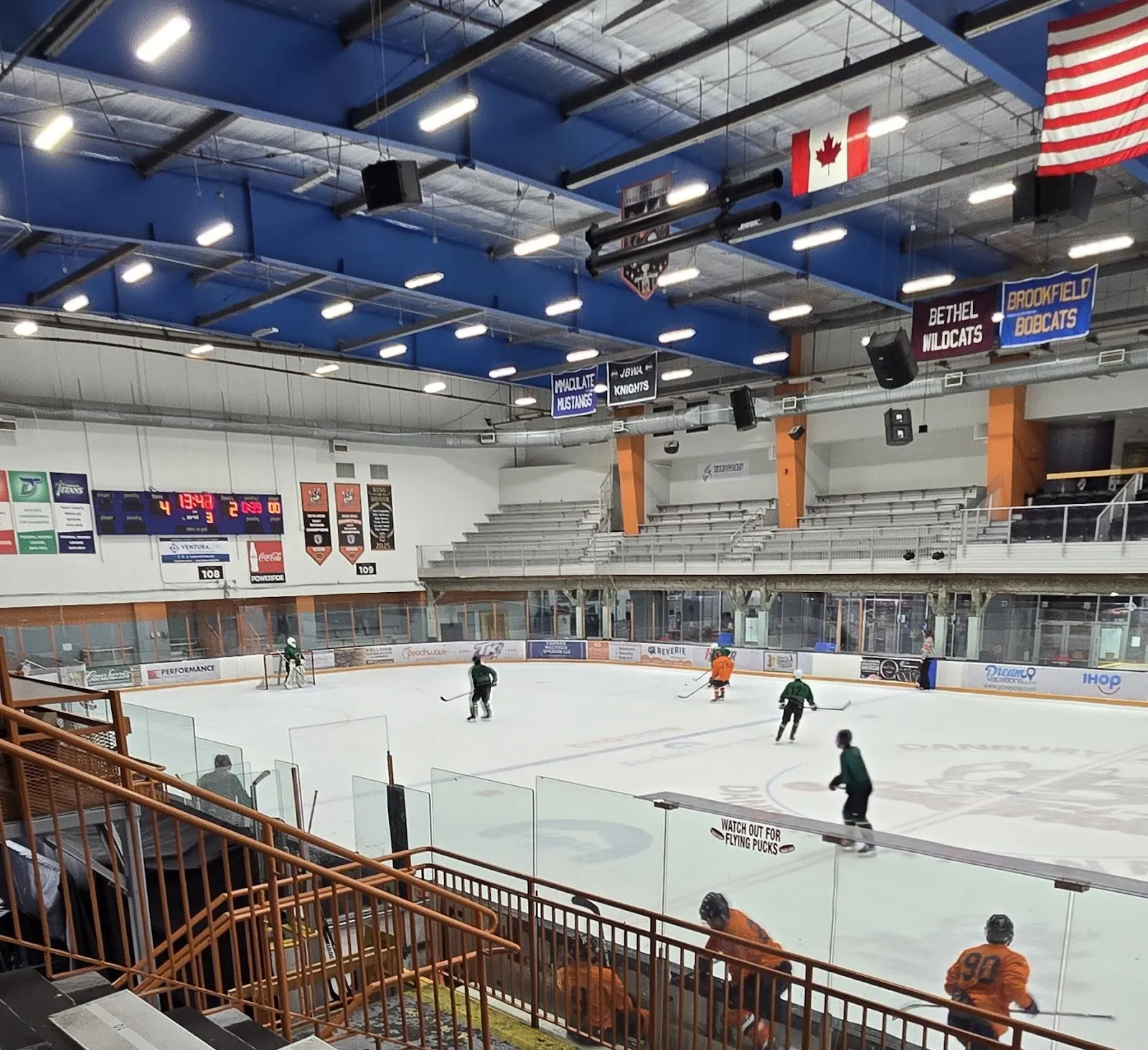 A game seen from the stands with a rail in front, players spread across the ice beneath a blue-trussed roof hung with Canadian and American flags. — Danbury Ice Arena
