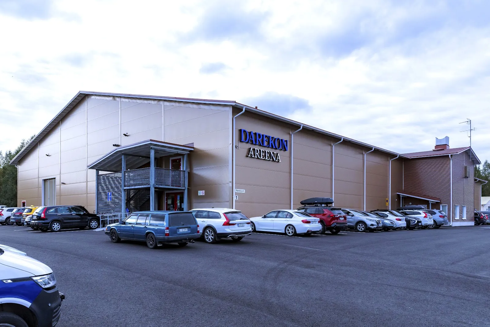 The pale blue-grey Haapavesi hall with a glazed entrance and cars parked along the front. – Darekon Areena