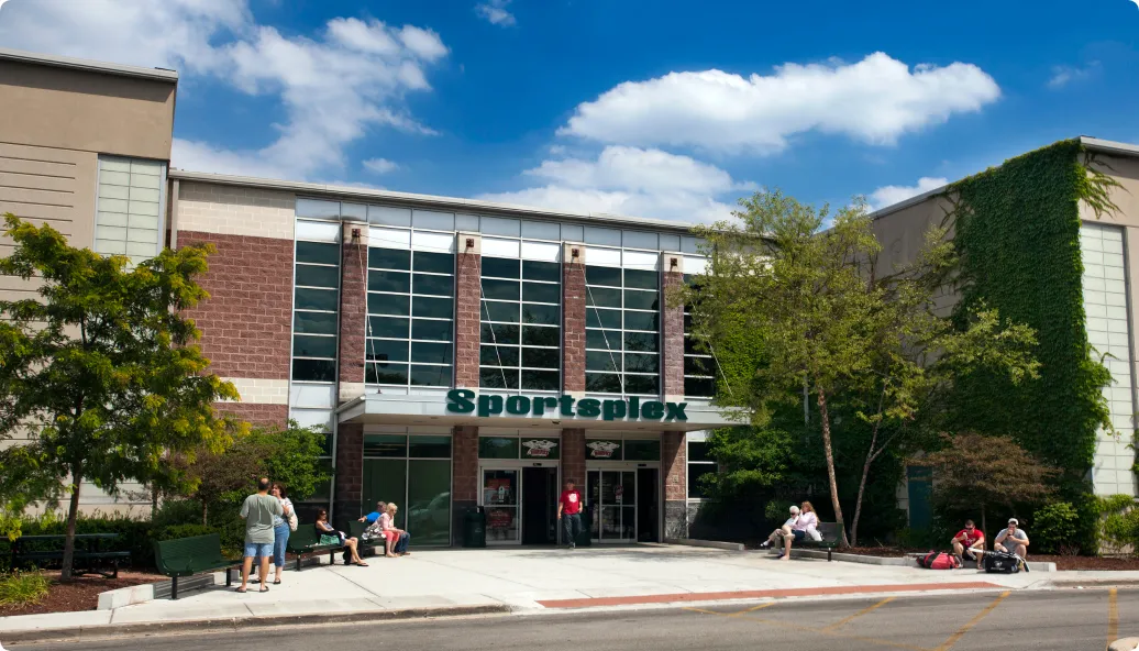 The Sportsplex front entrance on a sunny day: dark green 'Sportsplex' lettering on the canopy over the glass doors, a tall glazed brick window bay above, and benches on the plaza outside. — Darien Sportsplex
