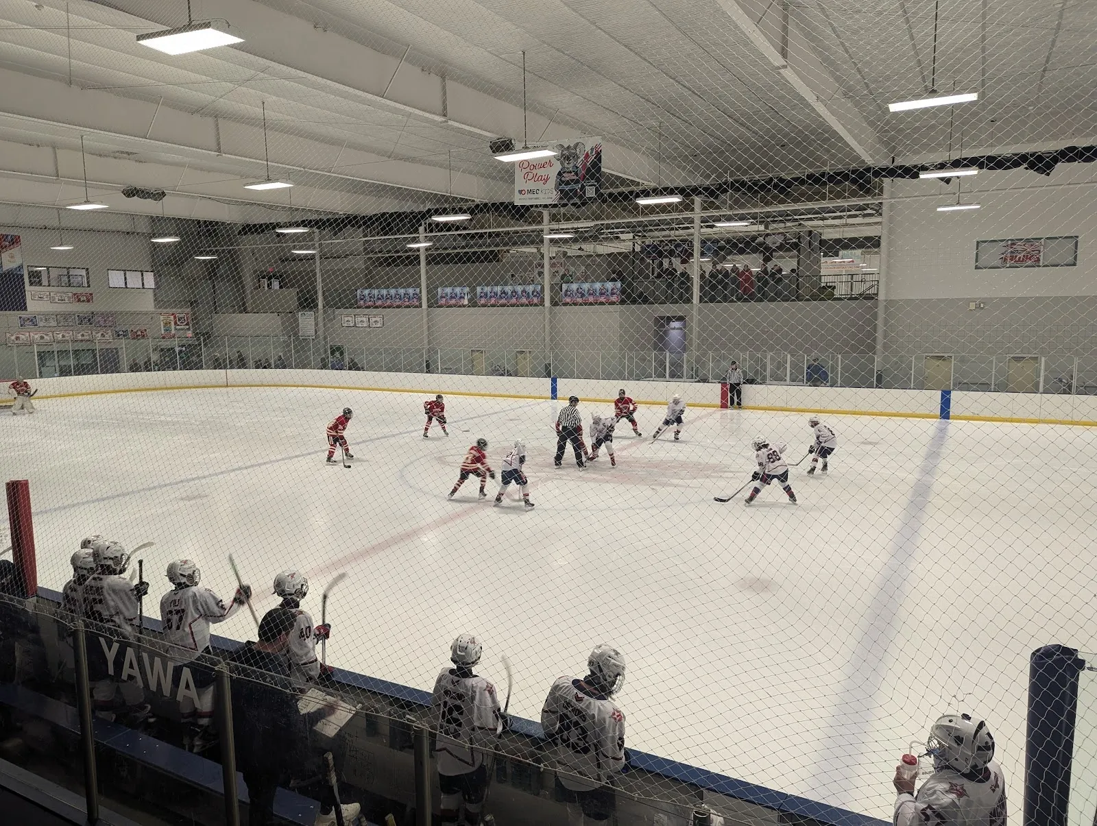 A wide view of the sheet from the stands, players spread across the ice with netting along the near side under a steel roof with flags on the end wall. — Darien Sportsplex