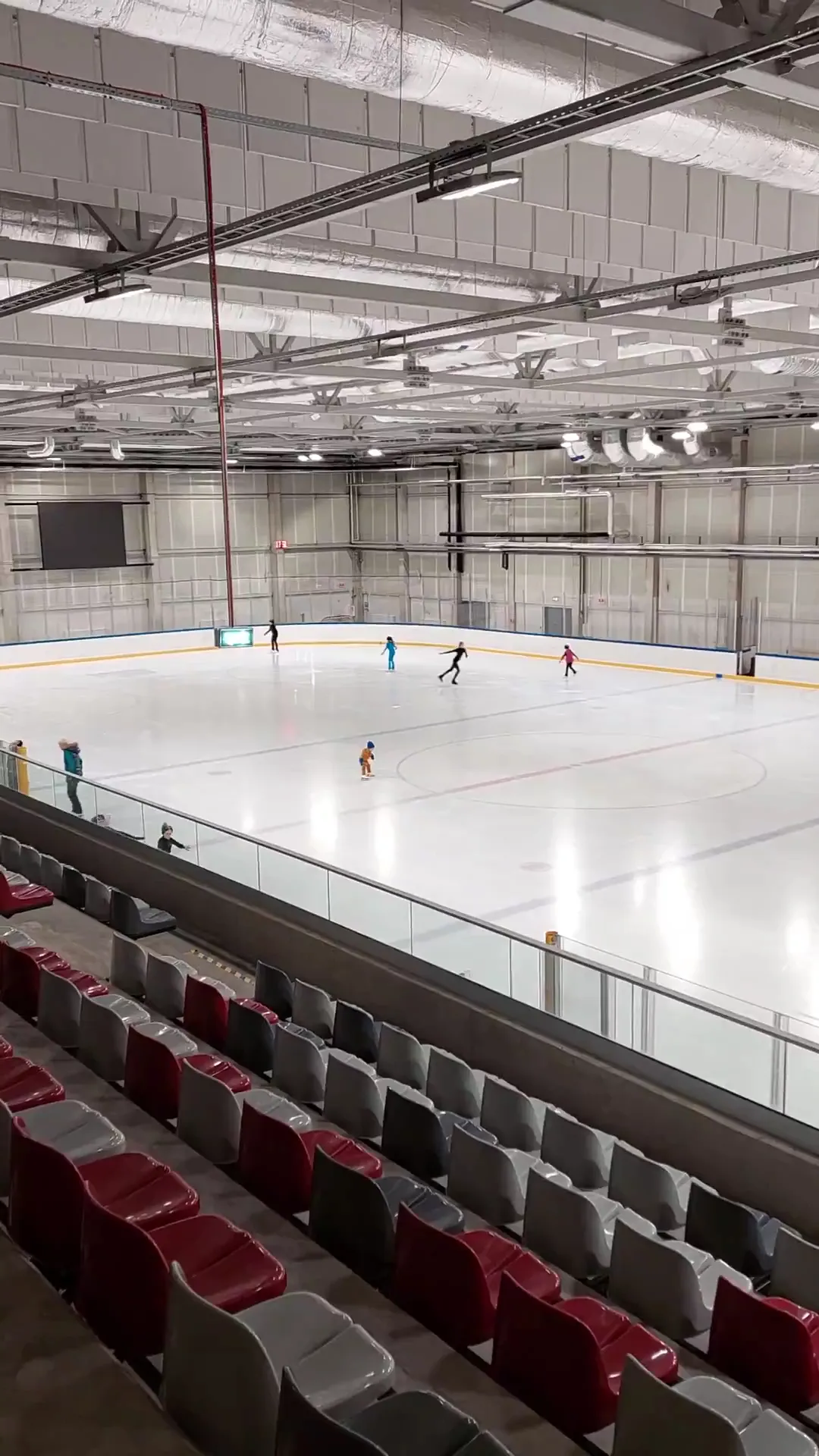 Ledus laukums Daugavas hallē, skatoties no tribīnēm: uz ledus slidotāji, priekšplānā sarkanu sēdekļu rinda. – Daugavas ledus halle