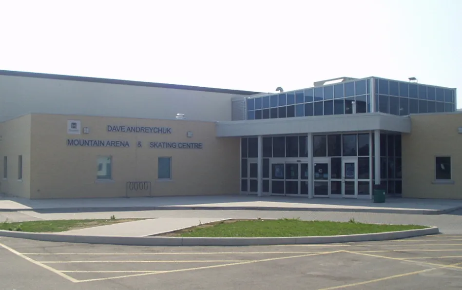 Entrance of the arena: a beige block with "Dave Andreychuk Mountain Arena & Skating Centre" lettering beside a glass lobby, a curb island and parking lines in front. — Dave Andreychuk Mountain Arena