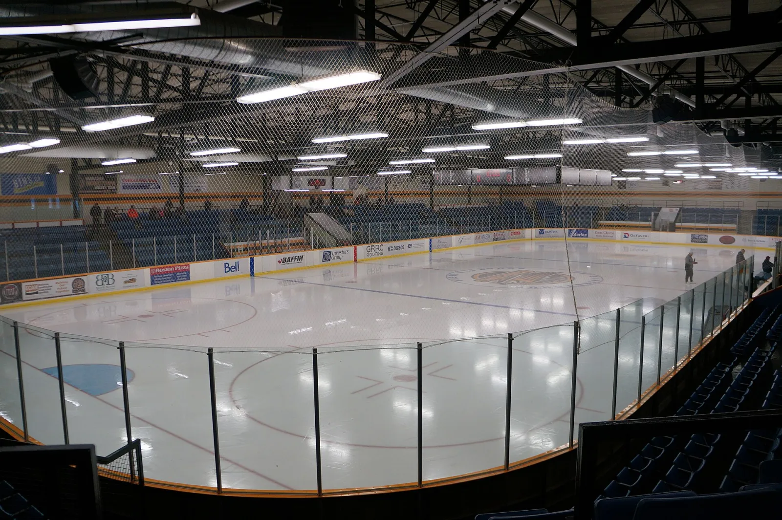 The empty main rink from the stands, with blue seats around the ice and a steel-truss roof. — Dave Andreychuk Mountain Arena
