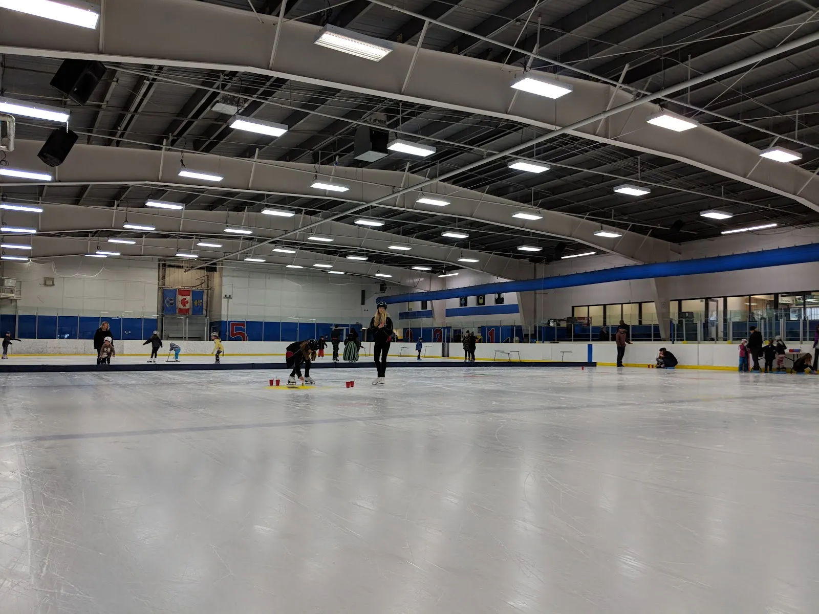 El hielo de Dave Barr bajo un techo de acero blanco, con algunos patinadores y una actividad infantil repartida en el extremo opuesto. — Dave Barr Community Centre