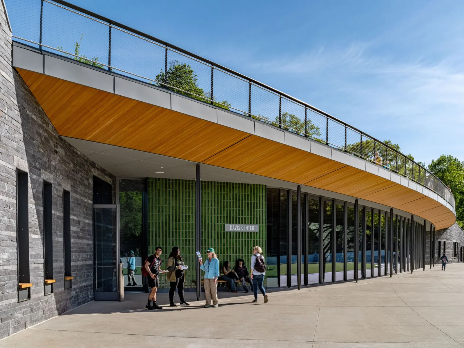 El edificio curvo de piedra oscura del Davis Center, con un toldo forrado de madera y puertas de vidrio a lo largo del andador, y el borde de un techo con plantas arriba. — Gottesman Rink at the Davis Center