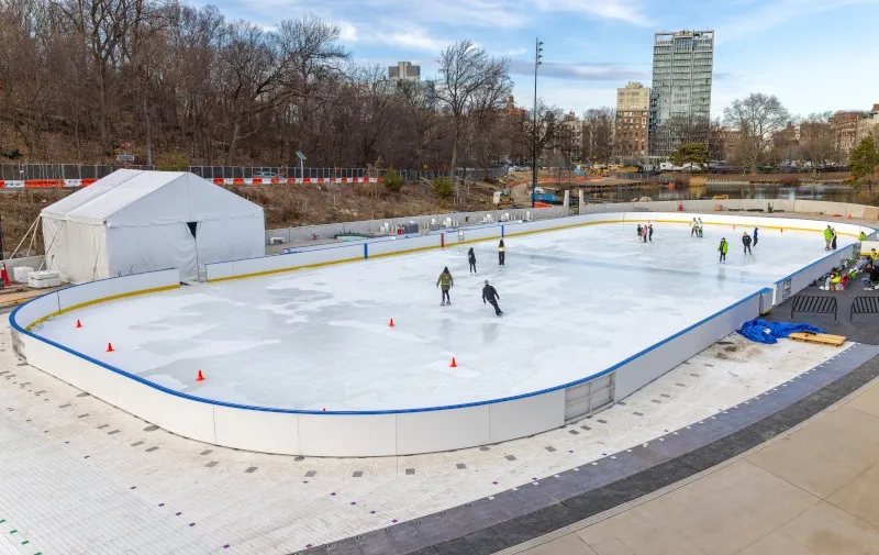 La pista ovalada Gottesman junto a una carpa blanca, con patinadores repartidos por el hielo y árboles sin hojas y torres del parque más allá de la valla. — Gottesman Rink at the Davis Center