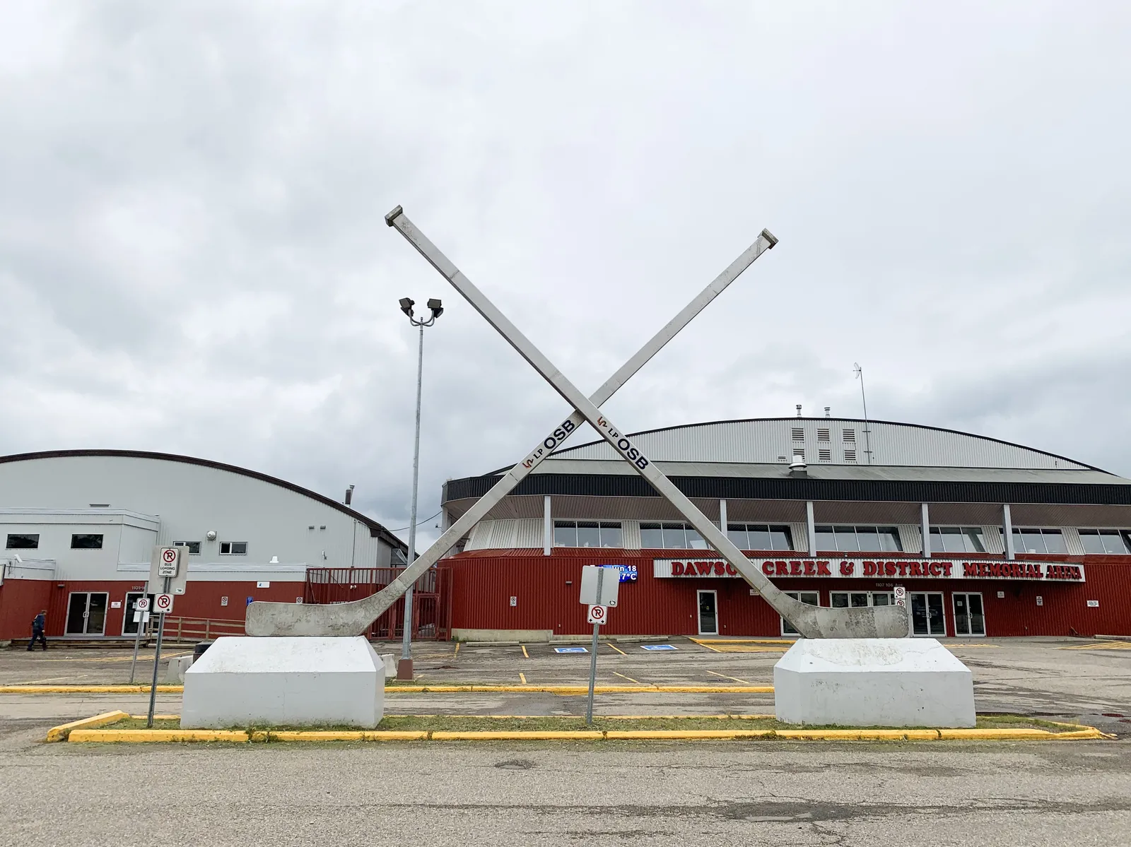 The Dawson Creek Memorial Arena seen across the car park under a grey sky, with a giant pair of crossed hockey sticks mounted in front of the building. — Dawson Creek & District Memorial Arena