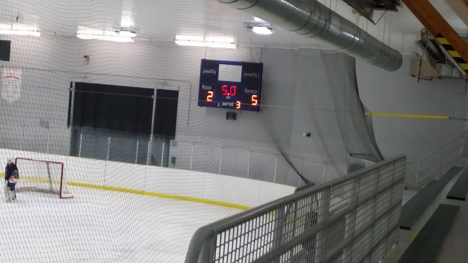The ice sheet at the Dawson Creek & District Memorial Arena seen from the stands, with the scoreboard, a goal at the far end and netting along the boards. — Dawson Creek & District Memorial Arena