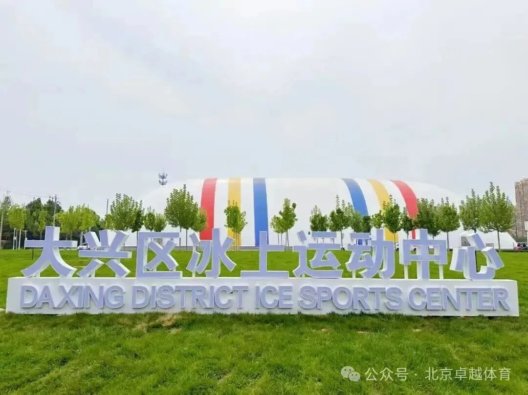 草坪上的大兴区冰上运动中心标牌，后方是覆盖冰场的彩条气膜馆 — Daxing District Ice Sports Center