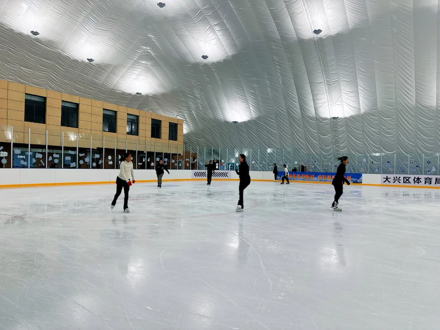 白色气膜下，滑冰者分散在冰面上，后方是界墙和两层的场馆用房 — Daxing District Ice Sports Center