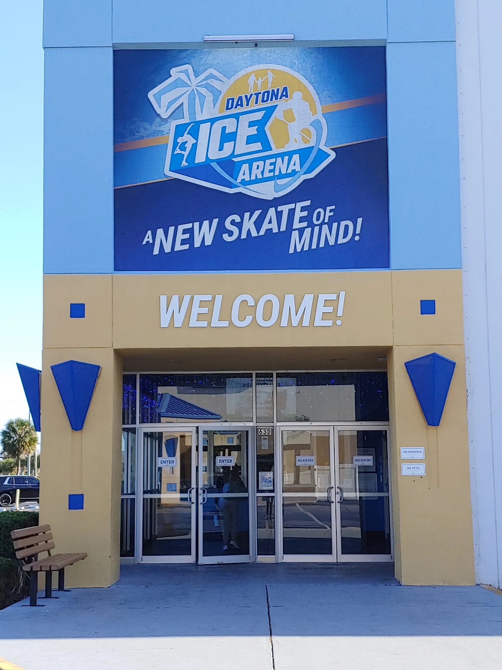 La entrada de Daytona Ice Arena, con una gran manta de A New Skate of Mind sobre las letras WELCOME y puertas de vidrio en una fachada beige y azul. — Daytona Ice Arena