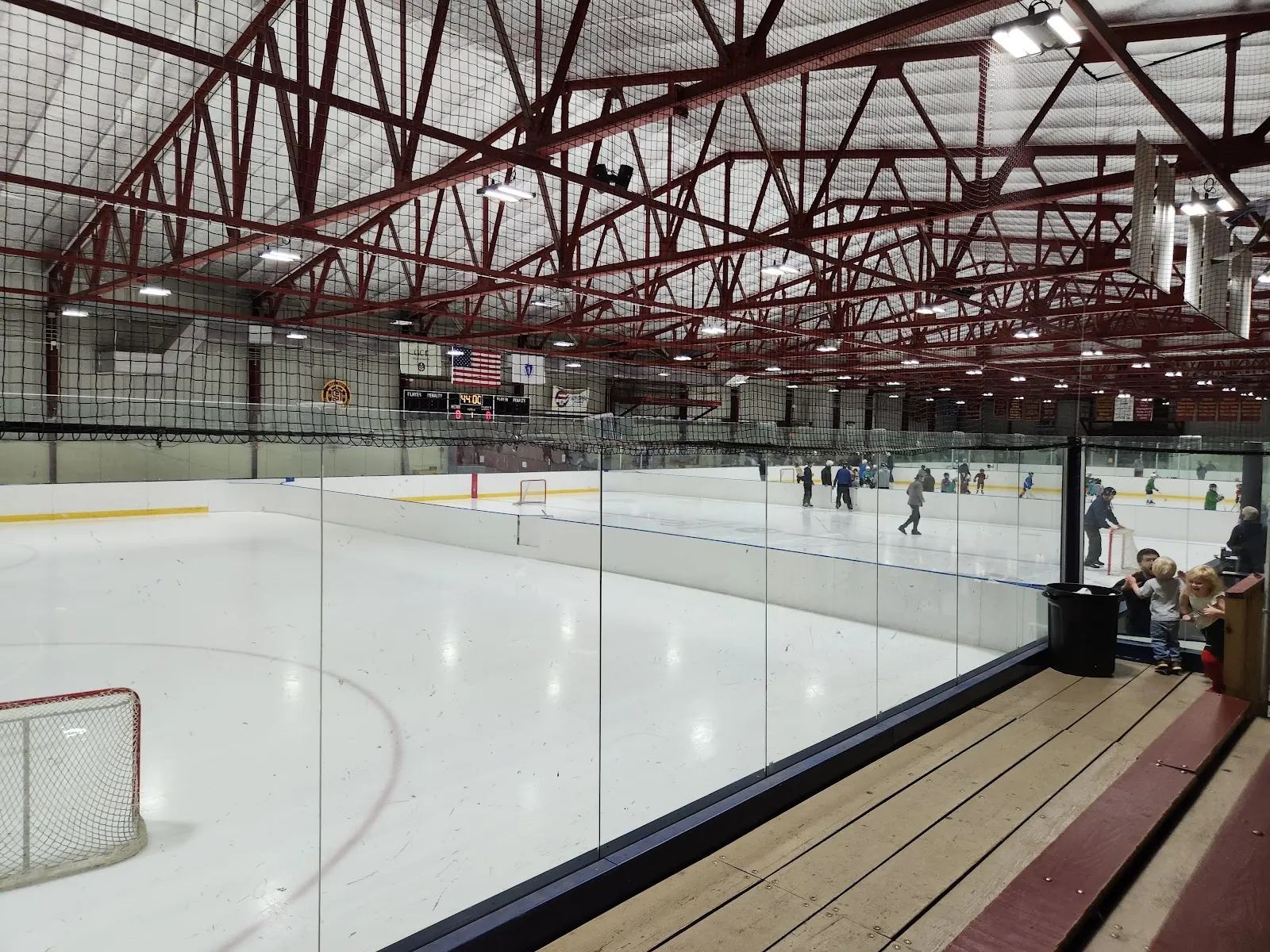 Amplia vista interior de las armaduras rojas de acero del techo, la pista y las vallas, con una sesión de patinaje público en curso en el lado opuesto del hielo. — DCR Bajko Memorial Rink
