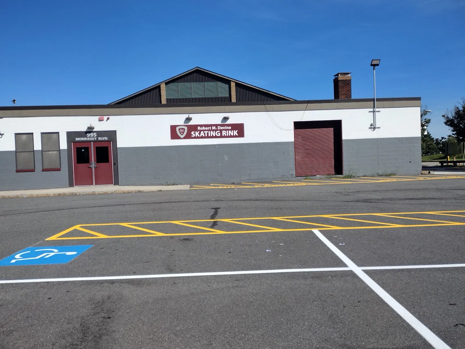 Front of the brick rink building at 995 Morrissey Blvd, with a maroon 'Robert M. Devine SKATING RINK' sign and DCR shield over the main entrance doors, empty parking lot in front. — DCR Devine Memorial Rink