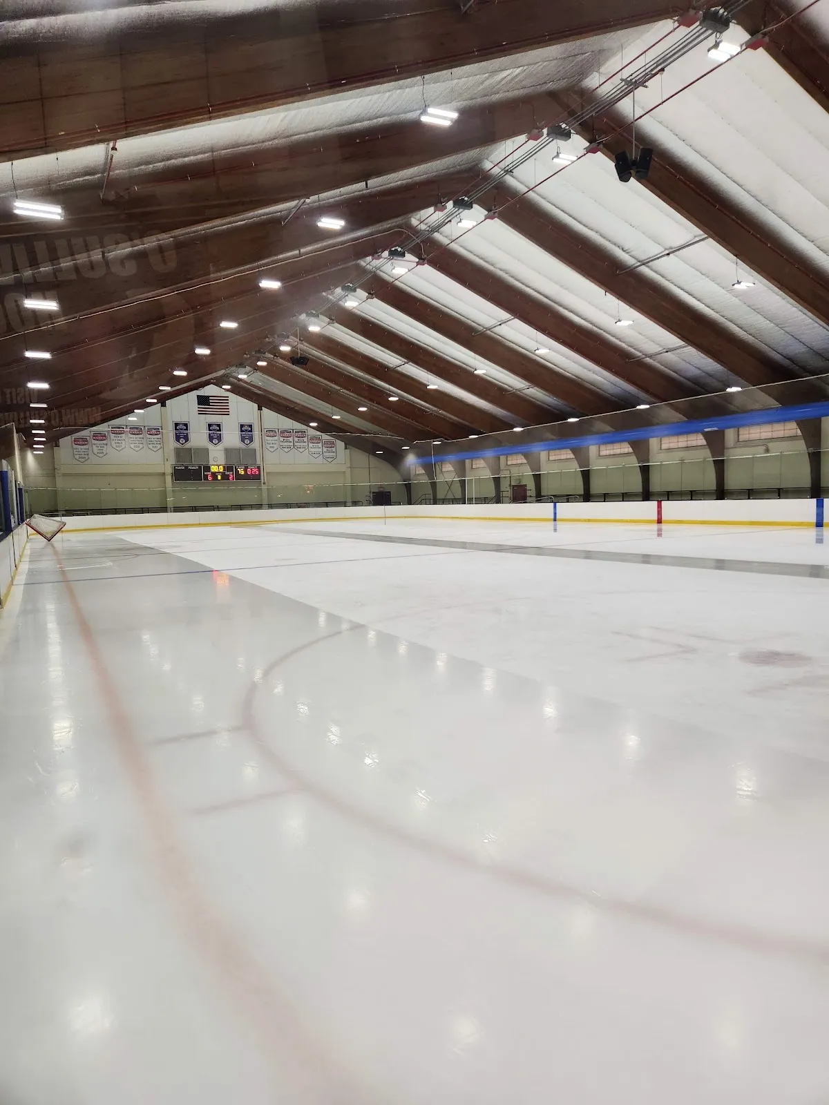 Empty ice sheet under a wood and steel vaulted roof, championship banners and an American flag on the end wall, no people on the ice. — DCR Devine Memorial Rink