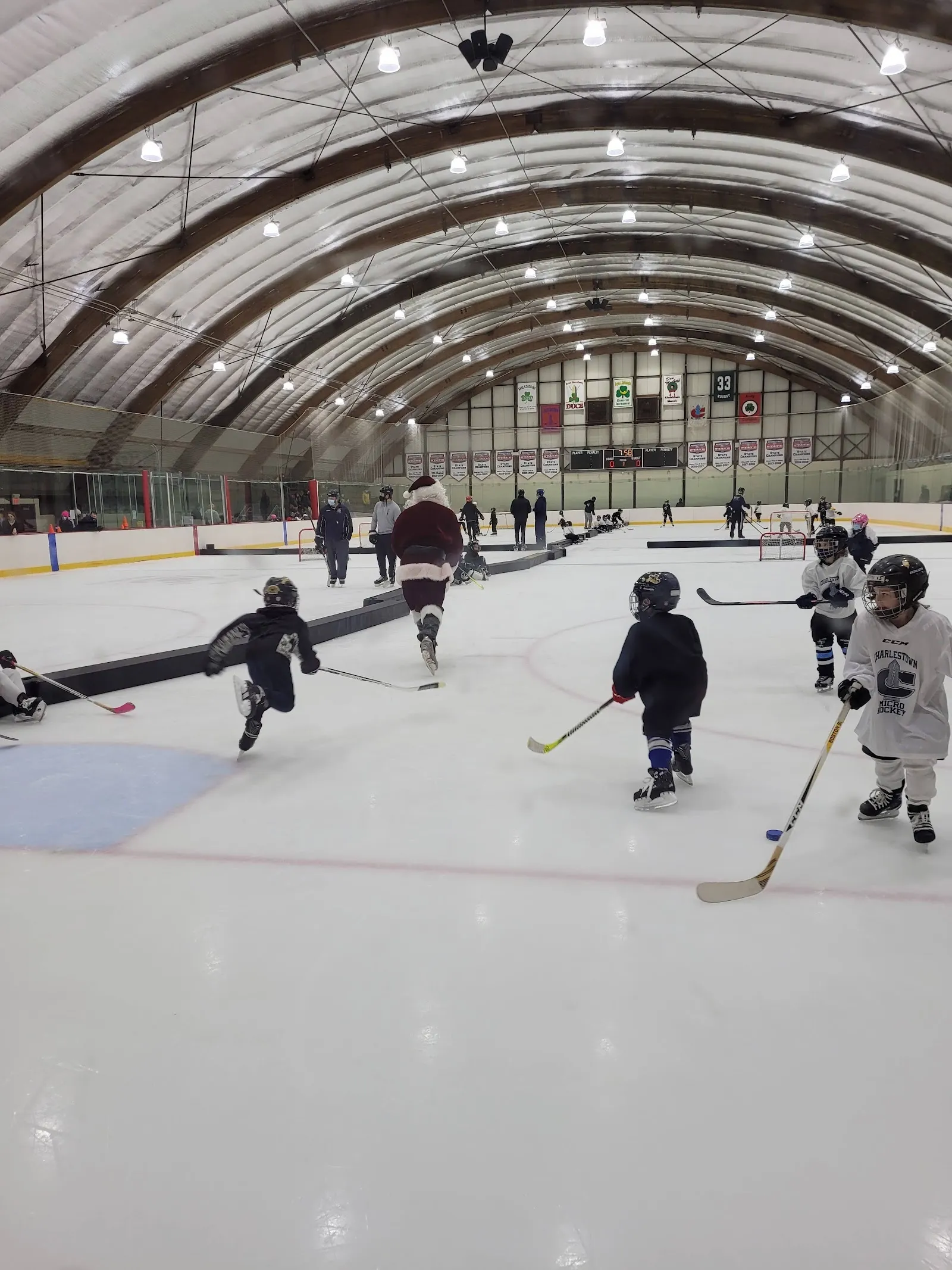 Jóvenes jugadores patinando a lo largo de las vallas cercanas bajo un largo techo arqueado, uno en primer plano y otros más adelante en la pista. — DCR Emmons Horrigan O'Neil Memorial Rink