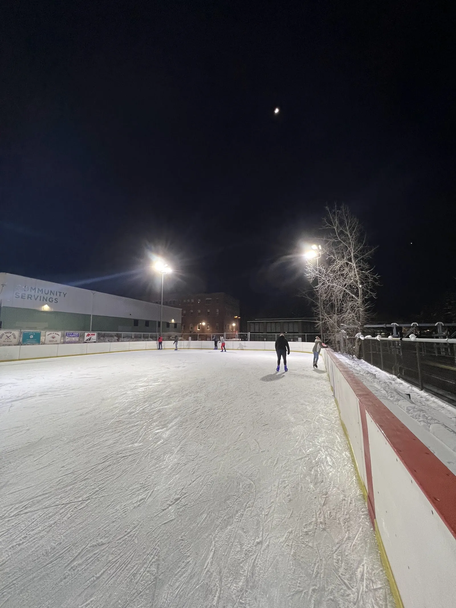 La pista al aire libre de Kelly bajo los reflectores de noche, mirando a lo largo de las vallas hacia el edificio de Community Servings en Amory Street — DCR Kelly Outdoor Rink