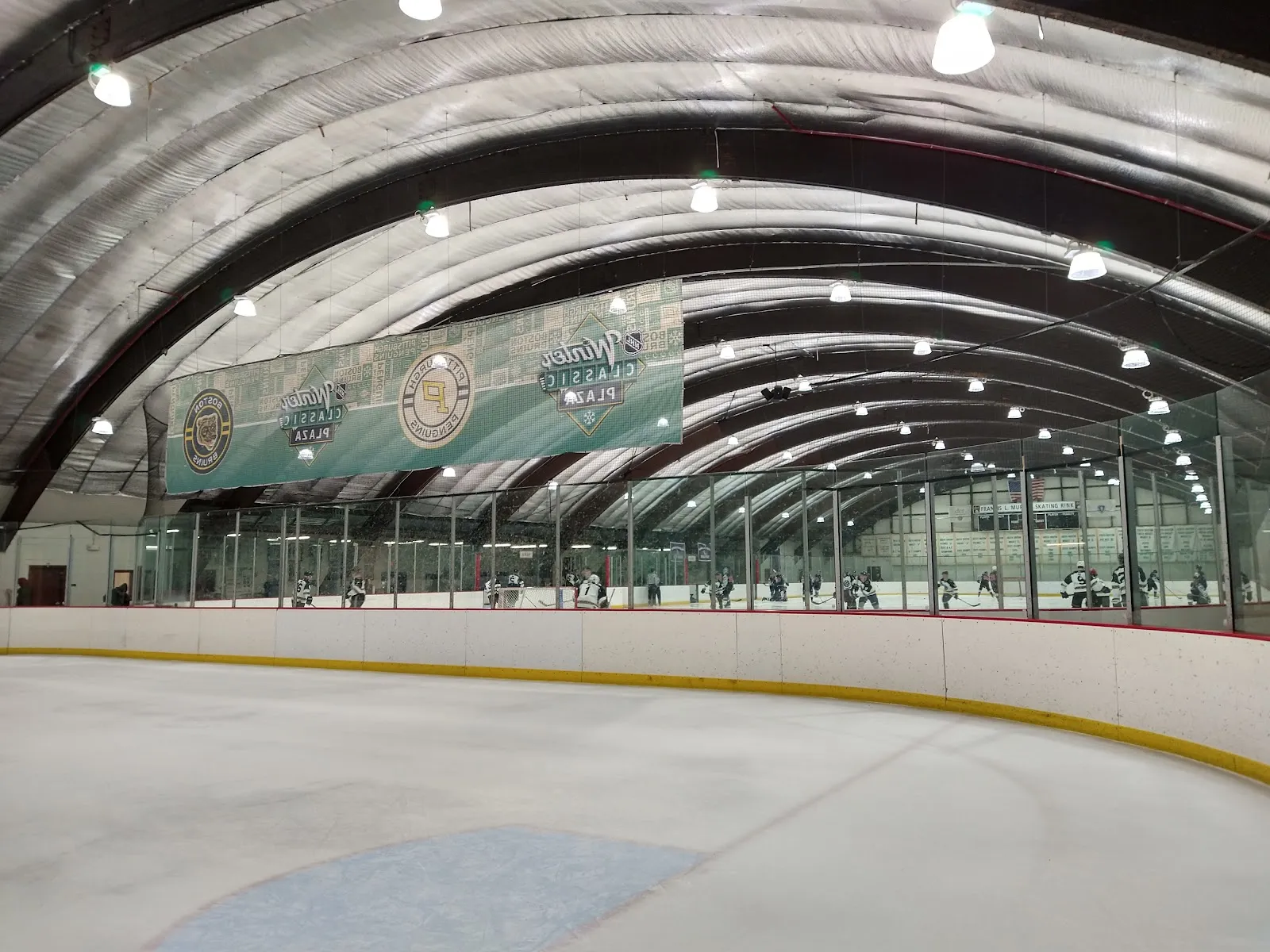 Amplia vista interior de la pista con techo de bóveda de cañón; sobre las vallas se ve un letrero en la pared que dice "FRANCIS L. MUR[PHY] SKATING RINK", con jugadores juveniles de hockey en un partido de práctica al fondo. — DCR Murphy Memorial Rink