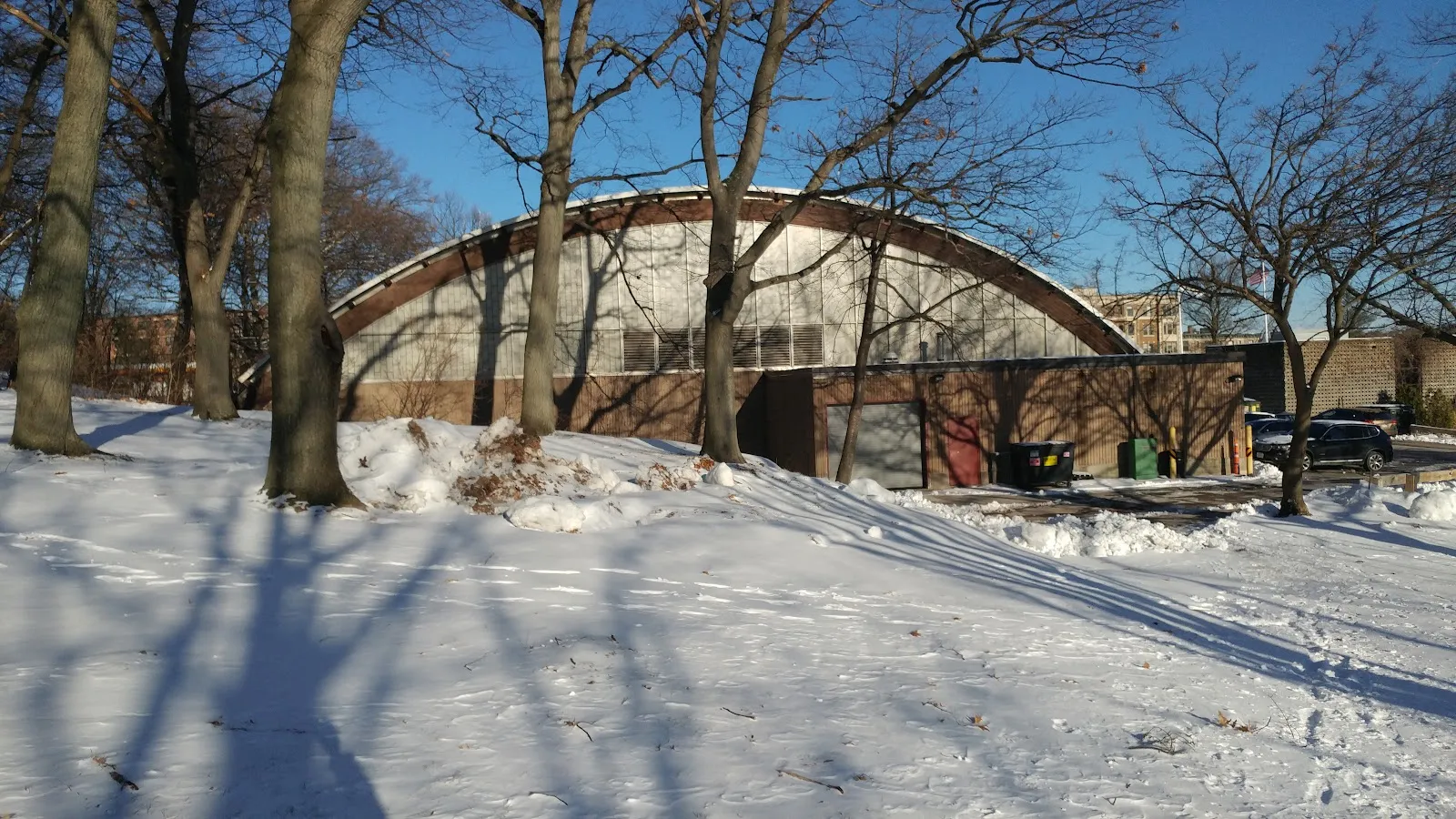 El techo abovedado en arco y la base de ladrillo de la Reilly Memorial Rink, vistos desde el otro lado de un césped nevado entre árboles invernales sin hojas. — DCR Reilly Memorial Rink