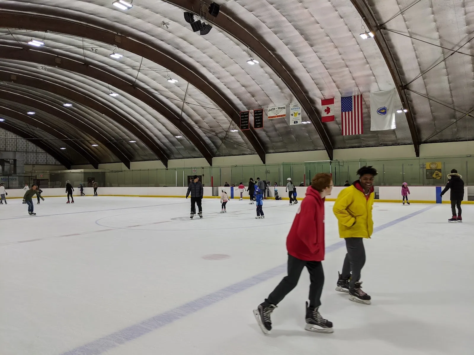 Una sesión pública bajo un techo de bóveda de cañón con banderas de Estados Unidos y Canadá, con patinadores repartidos por la pista y dos patinadores con chamarras brillantes en primer plano. — DCR Reilly Memorial Rink